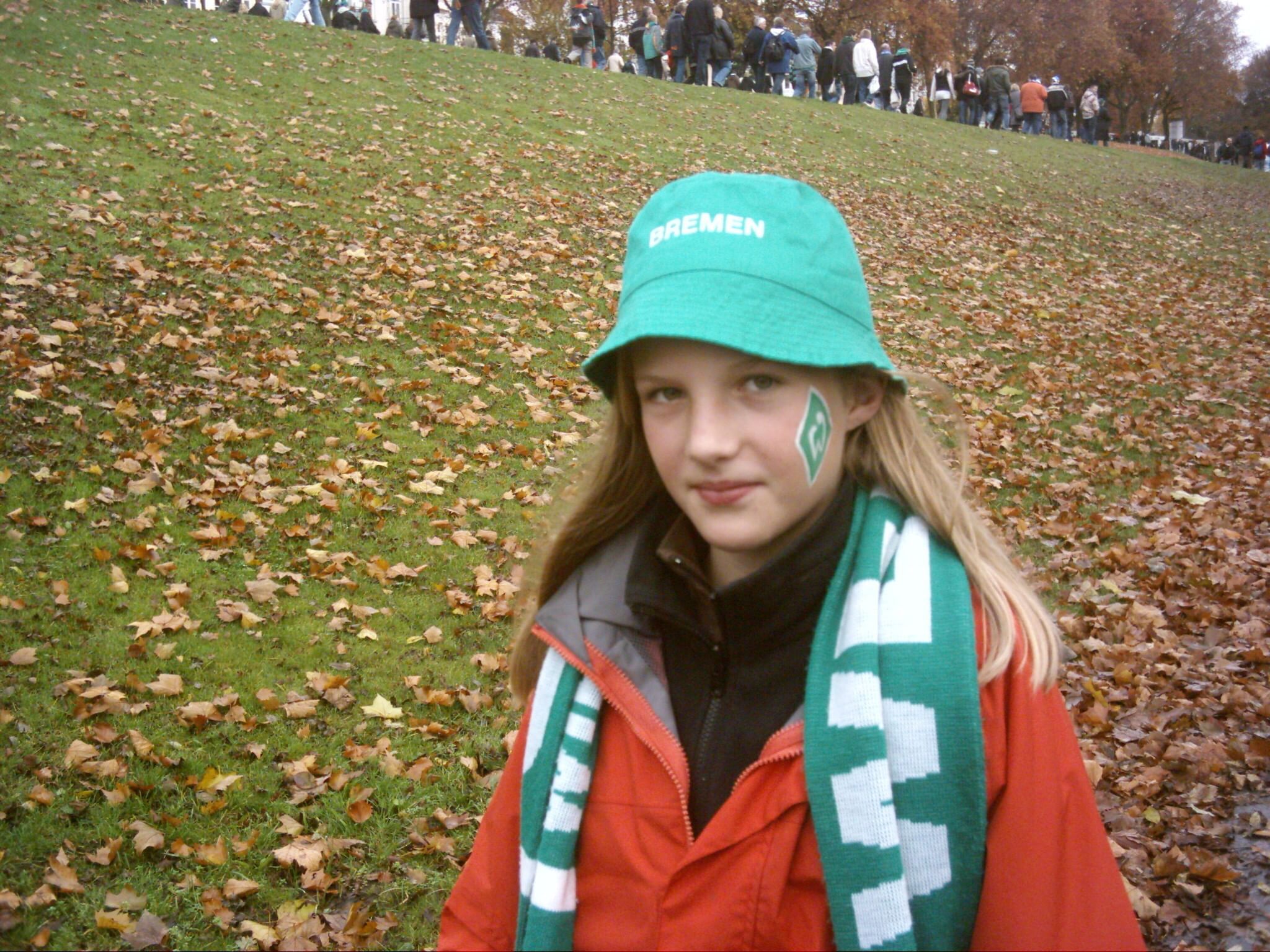 Lea Schönborn als Kind mit blonden Langen Haaren, einem Werderbrememn Hut und einem Werder Bremen Schal auf einer Wiese.
