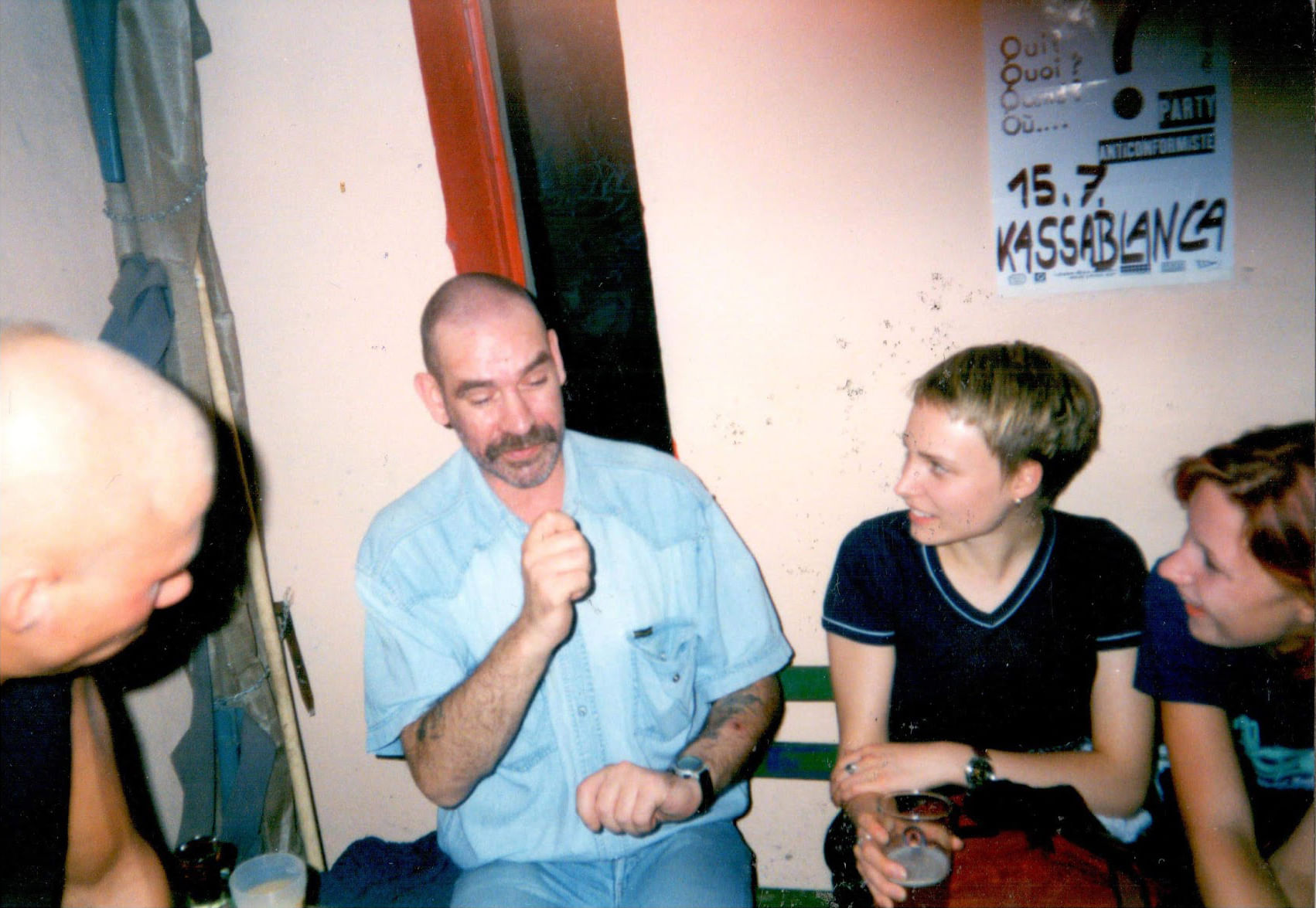 Analogfoto eines Mannes in seinen frühen 40ern, Kurzhaarfrisur und lichtem Haar in Richtung Halbglatze lehnend. Der Mann scheint die Umstehenden sehr gut zu unterhalten.
