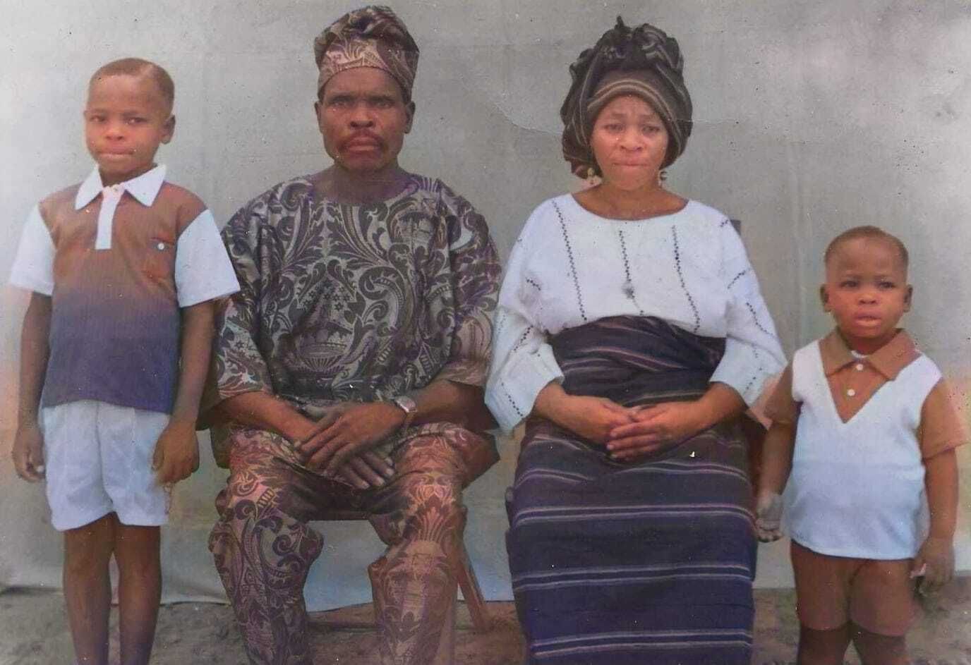 Ein coloriertes schwarz-weiß Bild, von links nach rechts: ein Junge, vermutlich im Alter um die 10 Jahre, ein Mann in typisch nigeriansicher Kleidung im Paisley Muster mit einer Art Mütze, eine Frau im Rock und weißer Bluse mit Kette und Kopfschmuck, ein Junge ca 5 im Pollunder.