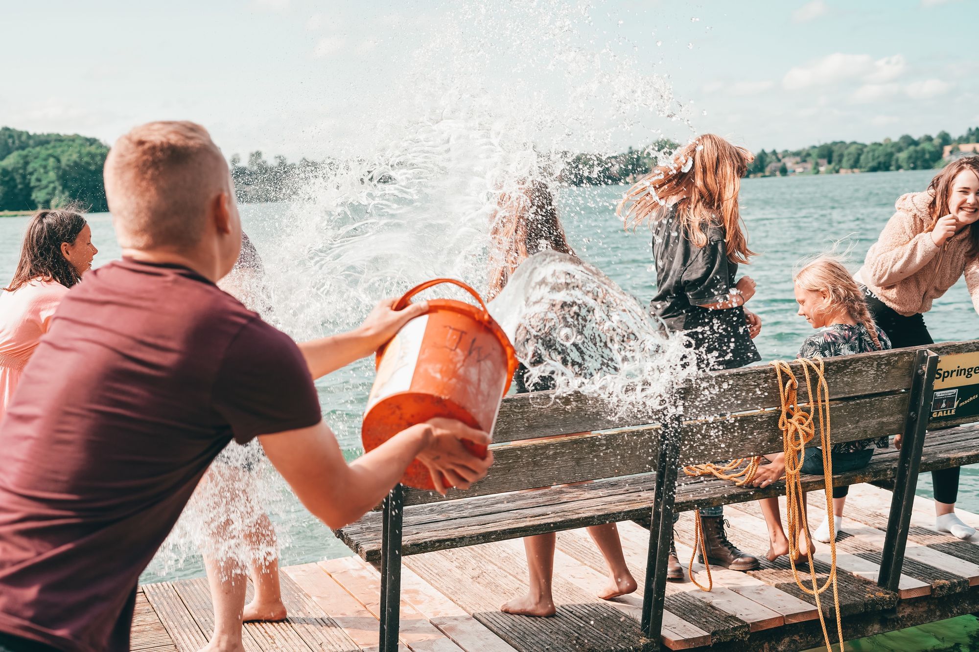 Ein Betreuer hat sich einen Eimer voll Wasser geschnappt und macht eine Gruppe von Mädchen auf dem Steg nass. 
