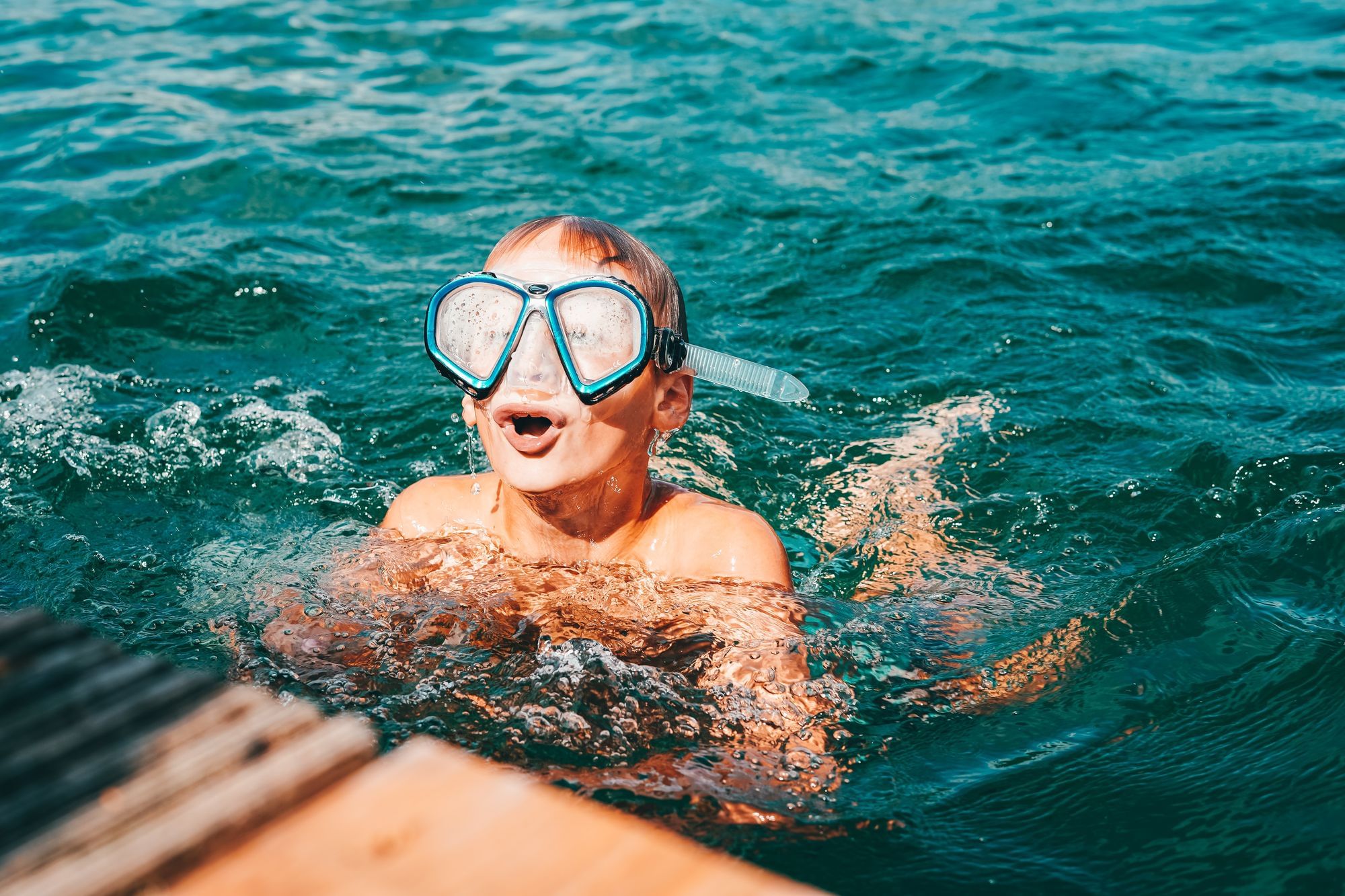 Ein Junge raucht mit einer Taucherbrille auf den Augen aus dem Seewasser auf und holt tief Luft.
