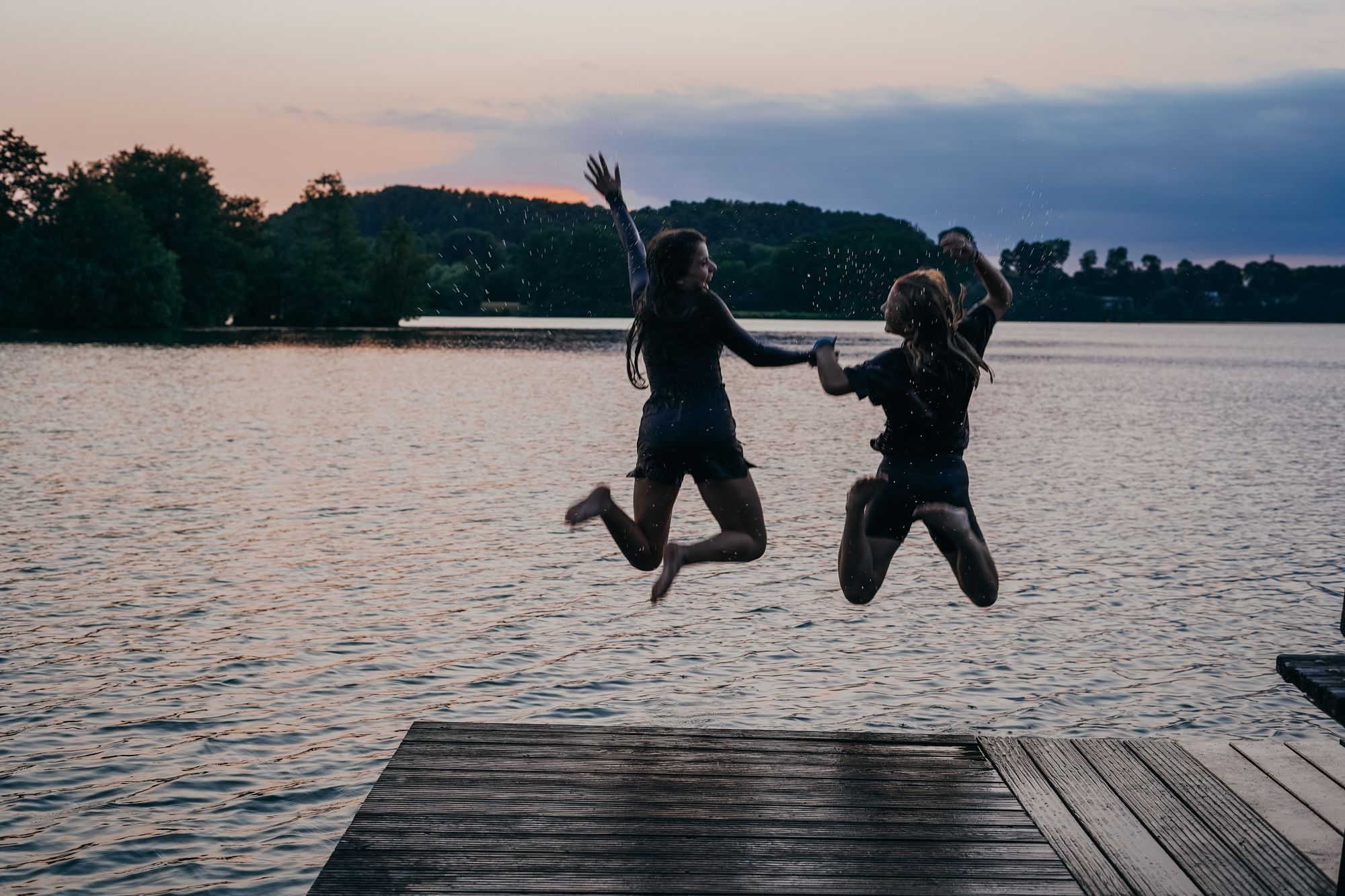 Zwei Mädchen springen Hand in Hand im Abendlicht in den Stocksee. 