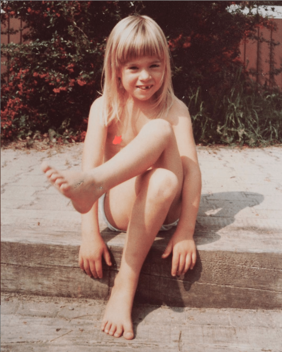 Ein ungefähr 6-jähirges Mädchen sitzt auf dem Boden und schaut mit übeinandergeschlagenen Beinen in die Kamera. Ein Sommerbild.