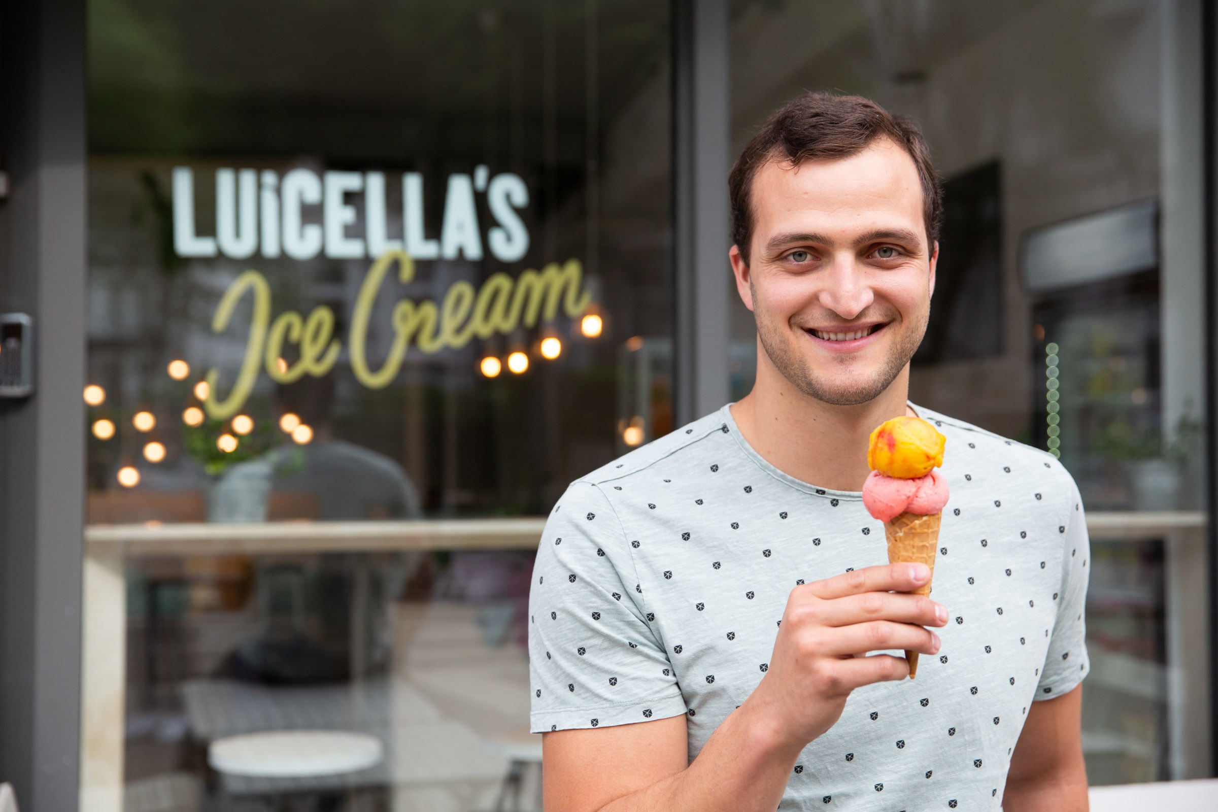 Wir sehen Markus Deibler, der ein Eis in der Hand hält und in die Kamera grinst – im Hintergrund sehen wir seinen Eisladen mit der Aufschrift Luicella's,