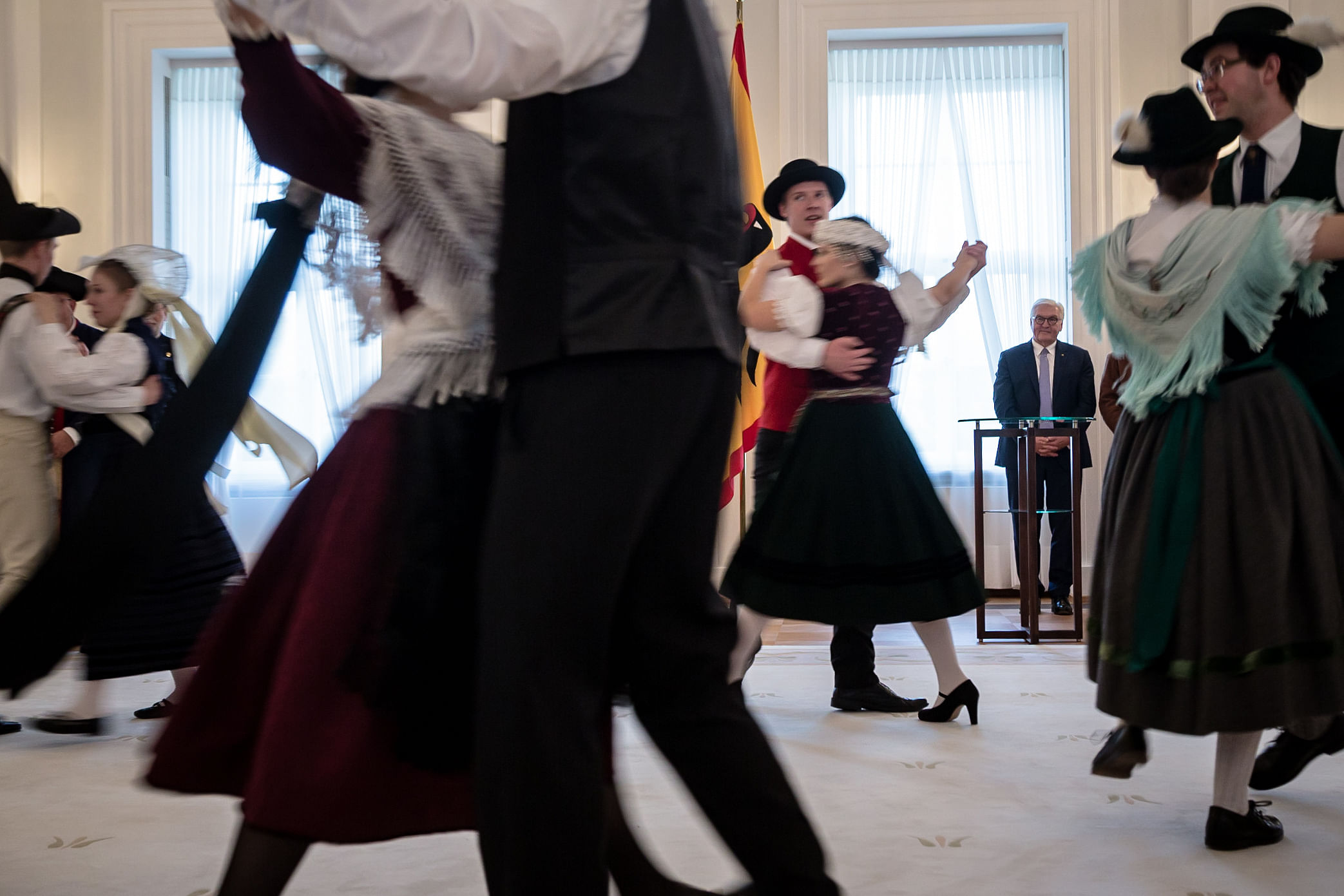Vier durch Bewegungsunschärfe nicht genauer erkennbare Trachtenpaare tanzen auf einem dicken Teppich im Schloss Bellevue. Frank Walter Steinmeier sieht ihnen zu, sein Gesichtsausdruck ist irgendwo zwischen freundlich und gequält.