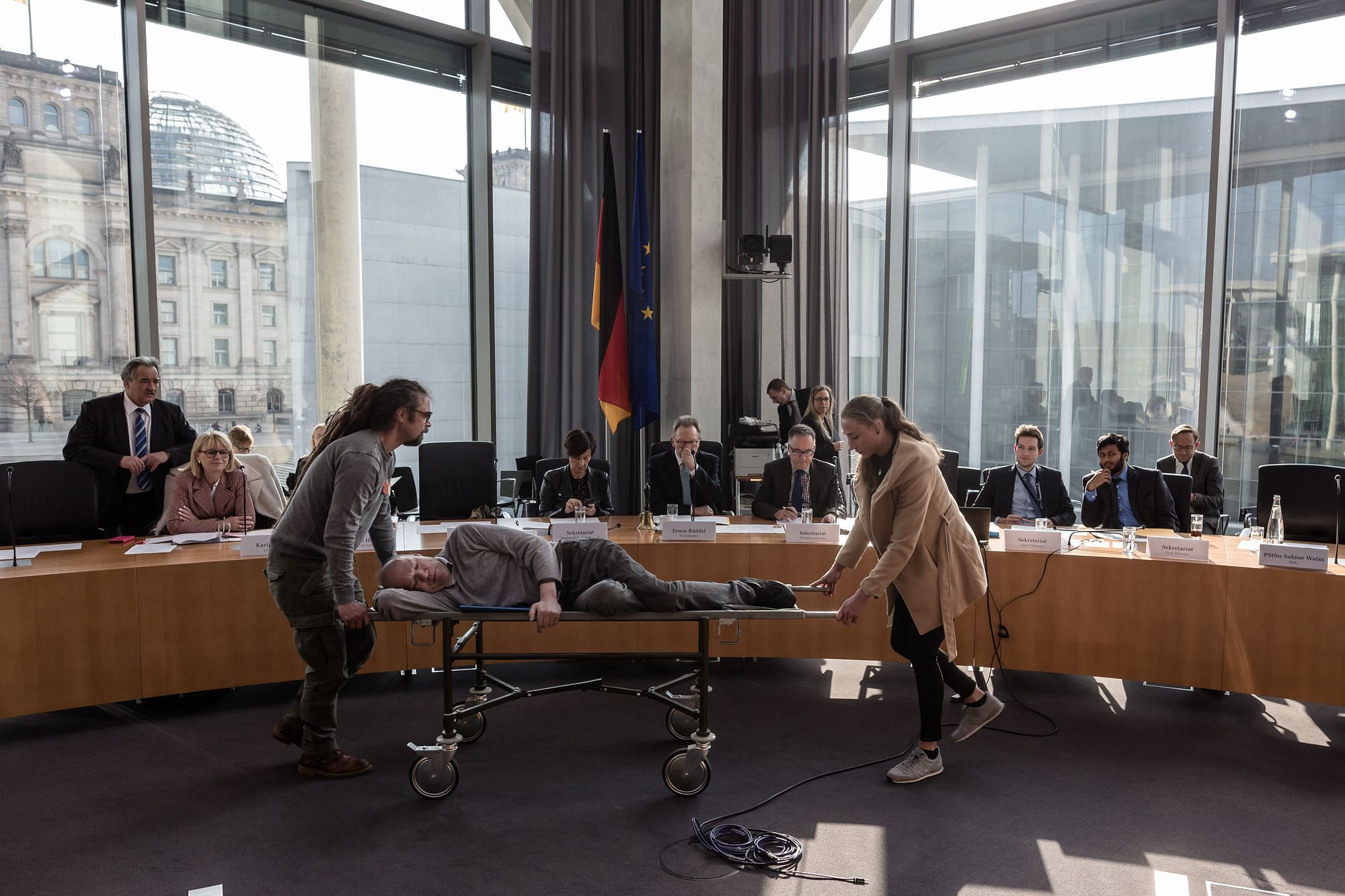 Anhörungssaal des Bundestag, wir sehen diesen im Hintergrund durch die Glaswand. Vor dem runden Tisch wird ein Mann von zwei Assistent:innen auf einer Bahre vorbeigeschoben.