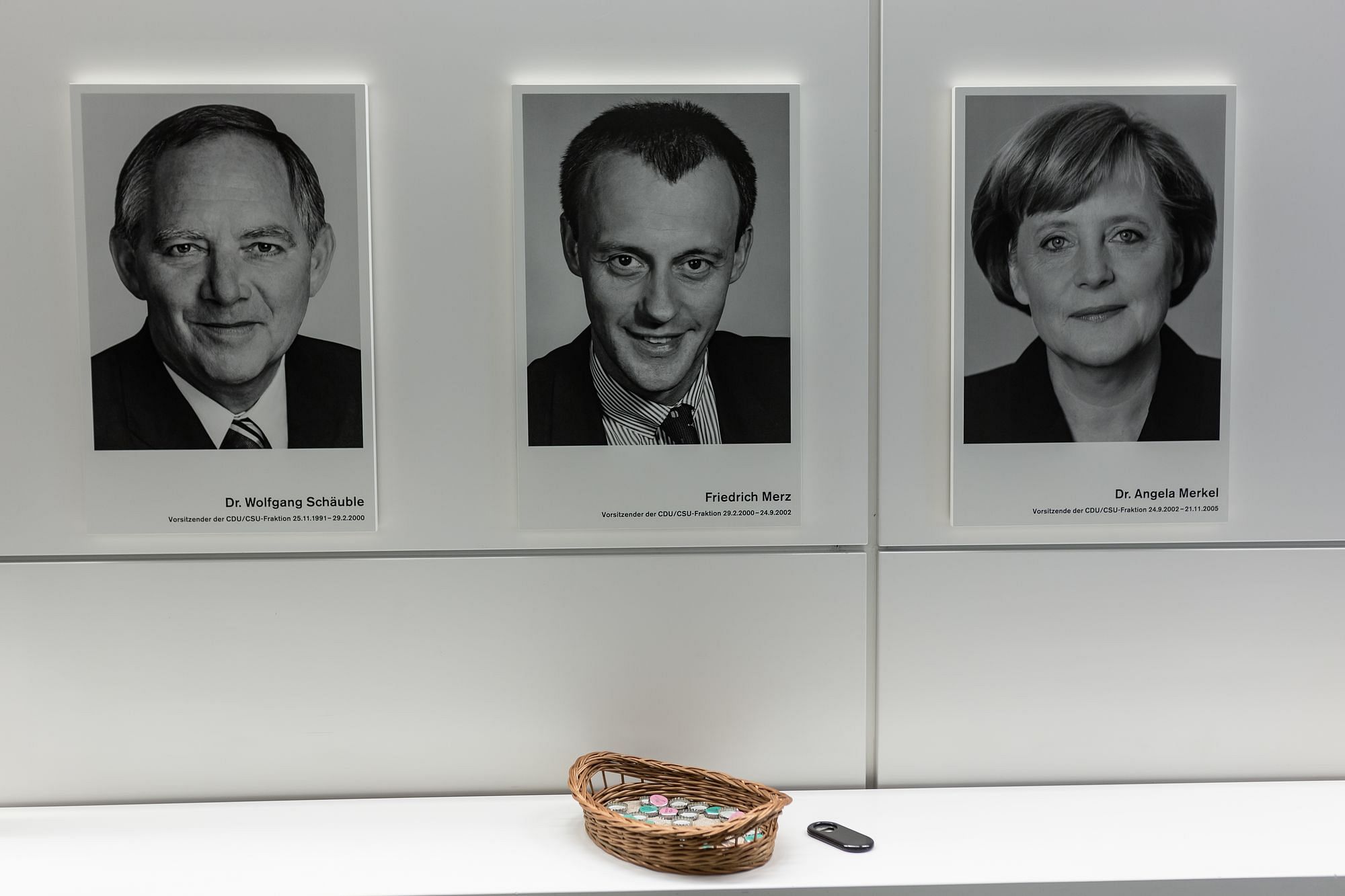 Eine weiße Wand mit Bildern. Von links nach rechts: Wolfgang Schäuble, Friedrich Merz und Angela Merkel (in jünger). Darunter ein Körbchen mit Kronkorken.