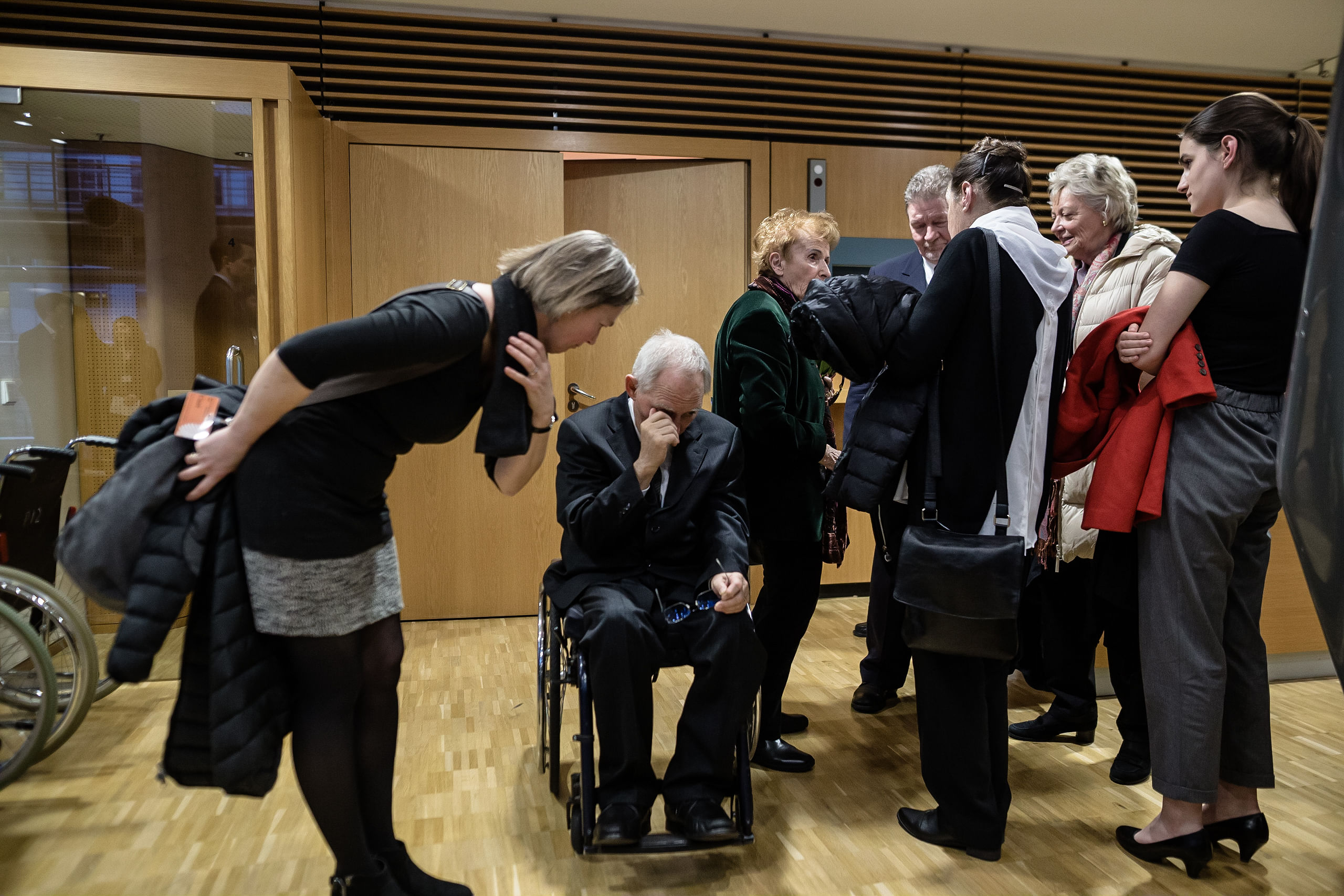 Wolfgang Schäuble vergräbt sein Gesicht in seinen Händen, hinter ihm steht eine Gruppe Menschen welche ihm keine Aufmerksamkeit bemisst, neben ihm lässt ihm eine weiblich gelesene Person Anteilnahme zukommen.