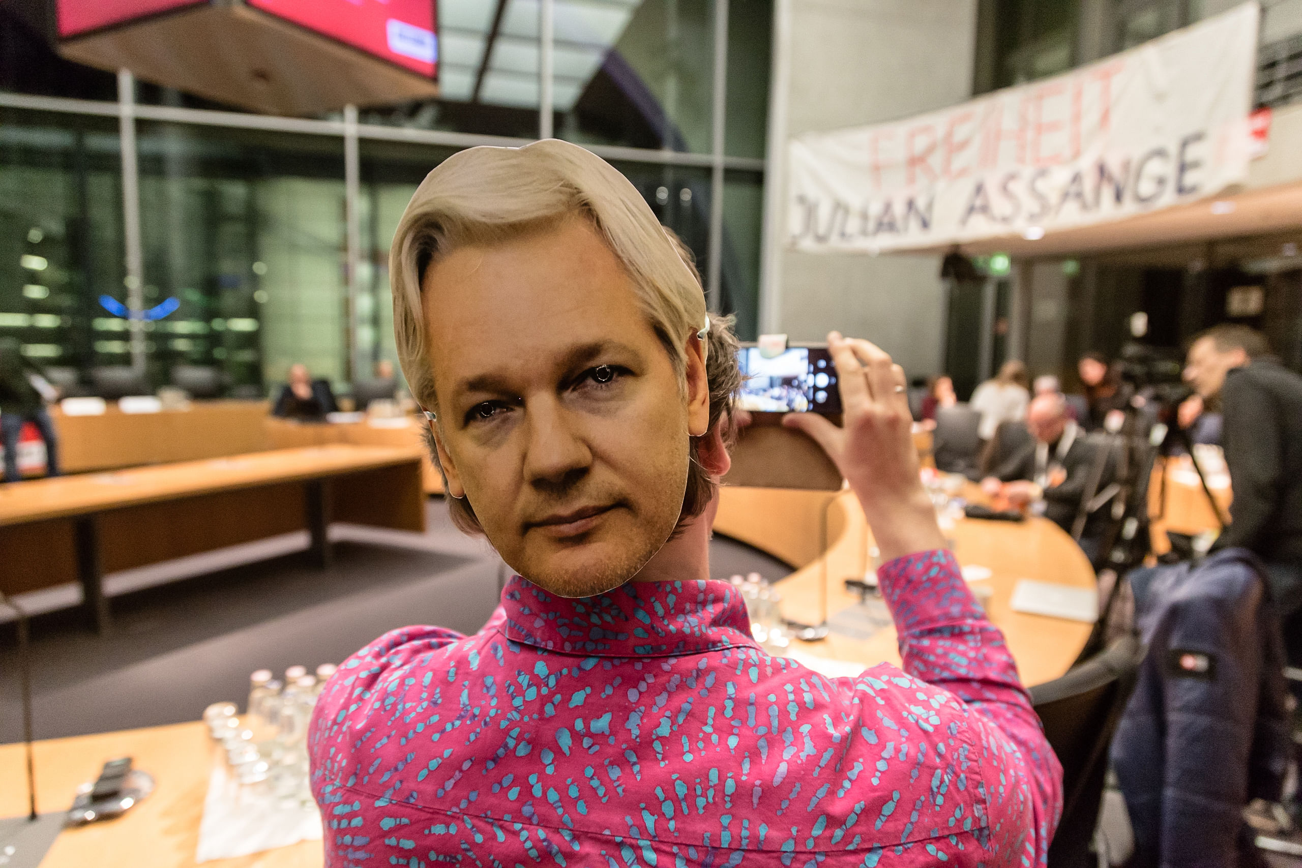 Anhörungssaal im Bundestag. Eine Person die uns den Rück zuwendet macht mit ihrem Handy ein Foto von einem Transparent auf welchem "Freiheit/ Julien Assange" steht. Die Person Trägt auf ihrem Hinterkopf eine Papierschnittmaske Assanges.