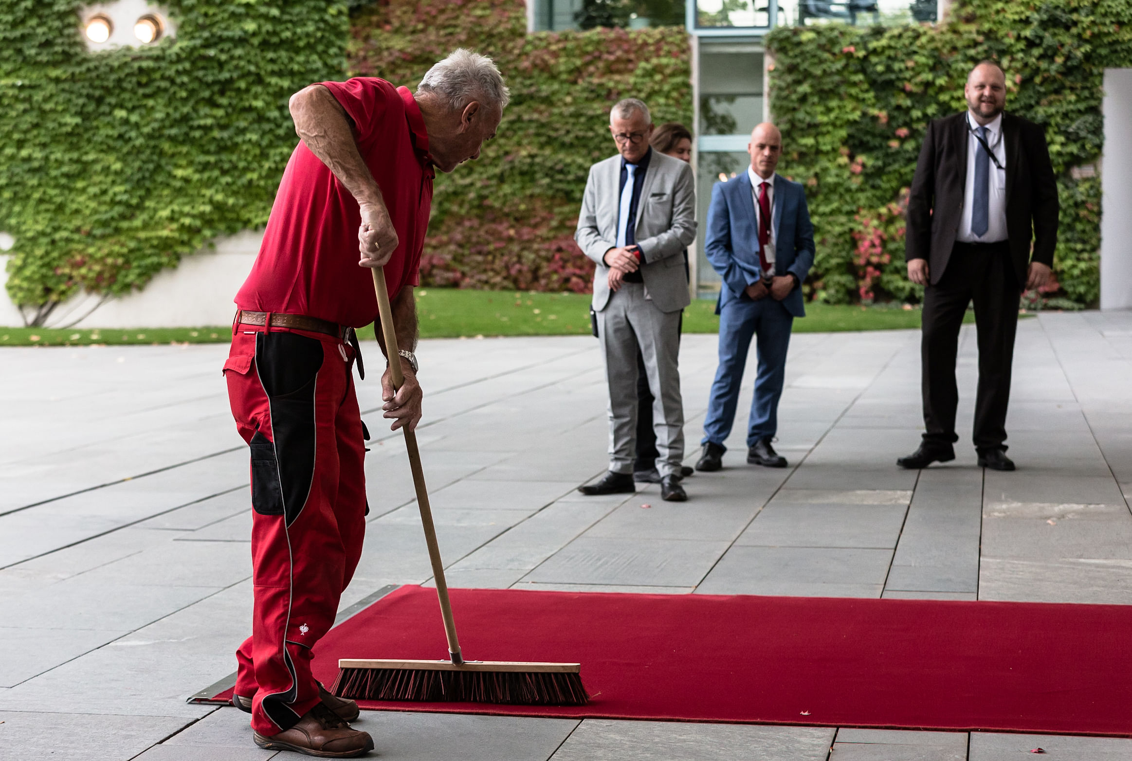 Ein rot gekleidter Hausmeister fegt den roten Teppich und wird dabei von drei Männer in Anzügen beobachtet.