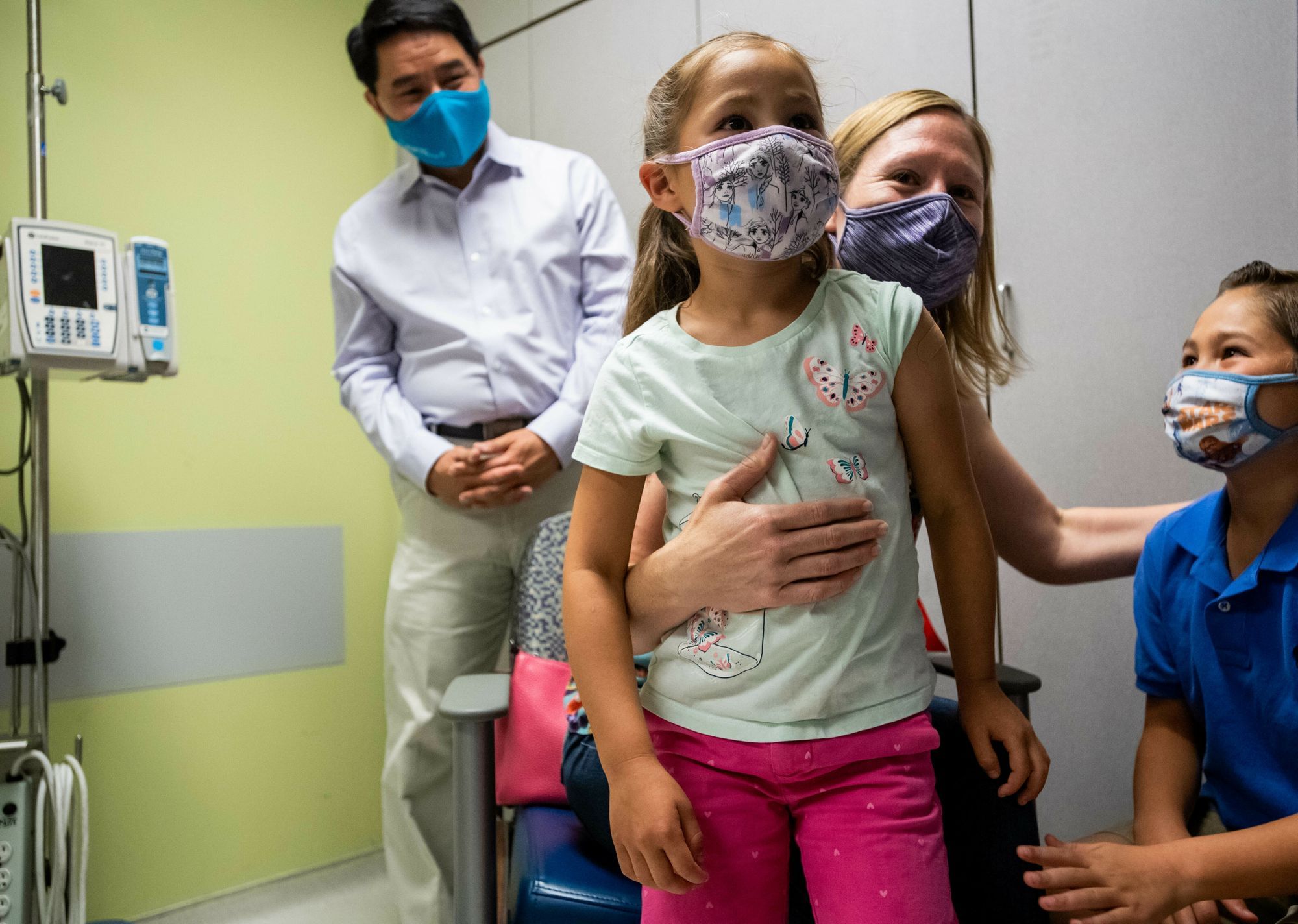 Krankenhausumgebung, alle tragen Mundschutz. Im Hintergrund ist ein Artz erkennbar. Eine Frau hält ein Kind vor sich, dieses steht, und ein anderes steht neben ihr, nur halb im