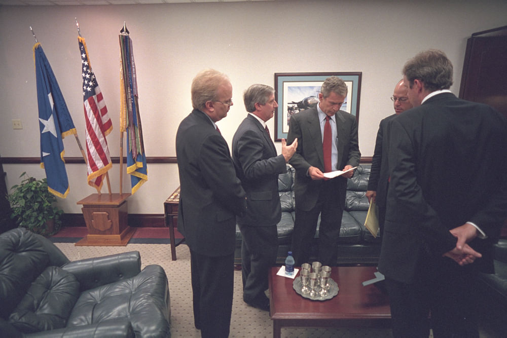 Eine Gruppe Männer, sie fokusieren sich auf den in einem Blattpapier vertieft. Es ist eine wohnlich-offizielle Atmosphäre, wir sehen drei Flaggen auf einem Ständer im Hintergrund. 