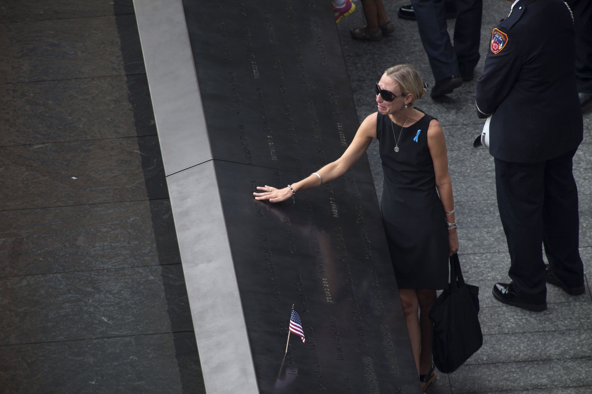 Teleaufnahme, eine in schwarz gekleidete Frau mit Sonnenbrille schluchzt während sie einen eingravierten Namen auf dem World Trade Memorial nachfährt.