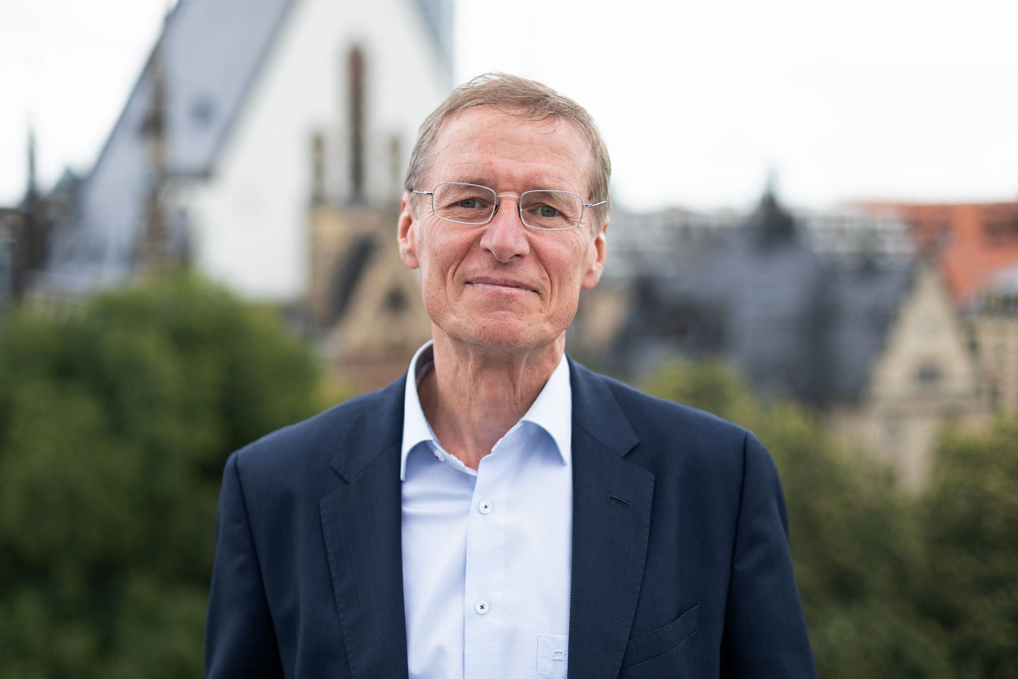 Ein Mann mittleren Alters mit Brille mit Hemd und Sacko, ohne Krawatte und vor einer verschwommenen urbanen Landschaft.