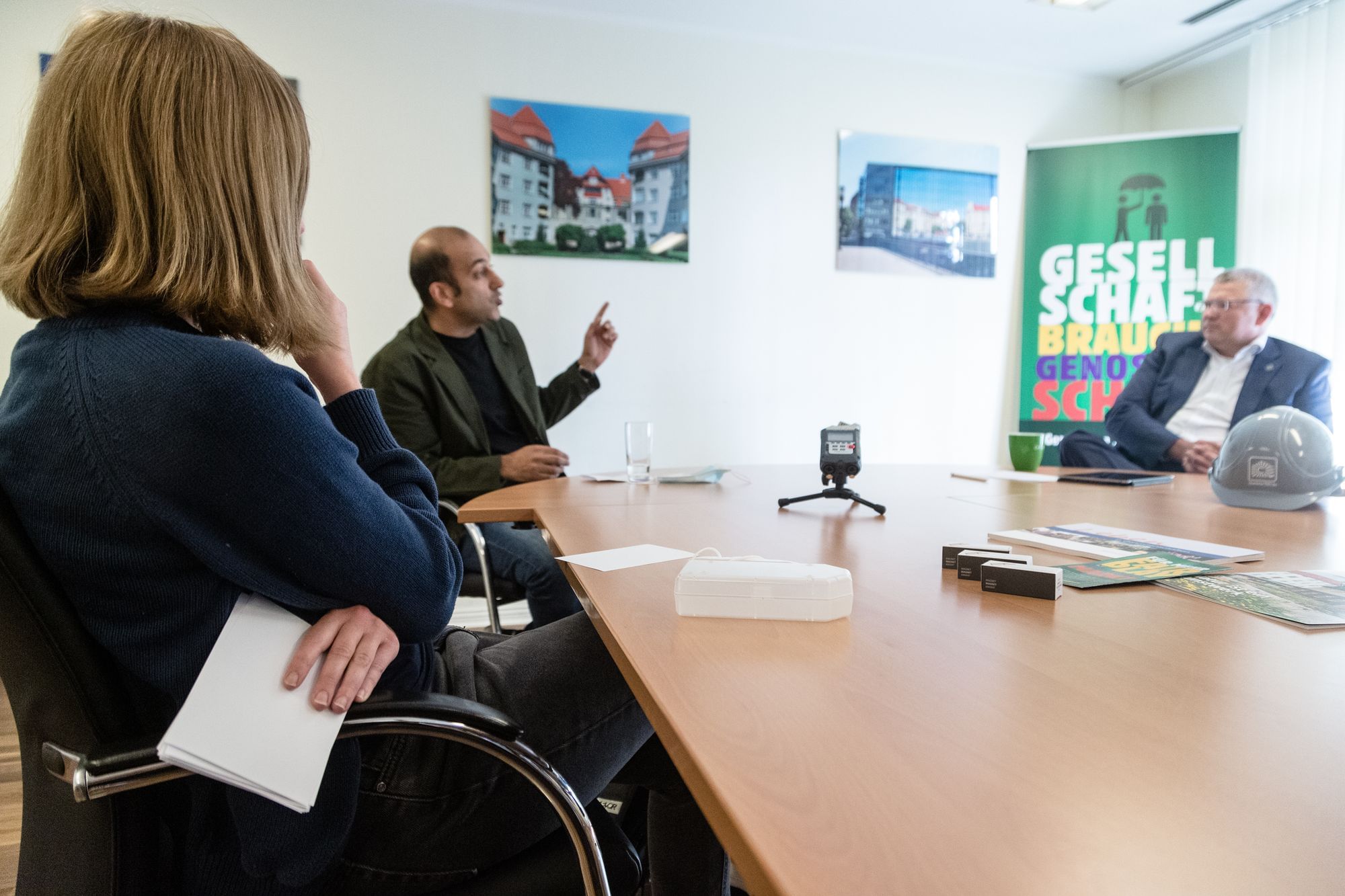 V.n.l.r.Rebecca Kelber dreht uns den Rücken zu, Karten in der einen Hand und die andere in nachdenklicher Pose, dahinter Herr Taheri der gerade argumentiert und Herr Enzensberger der aufmerksam zuhört. Der Raum ist mit Immobilienfotografie und Progesellschafts Werbung dekoriert.