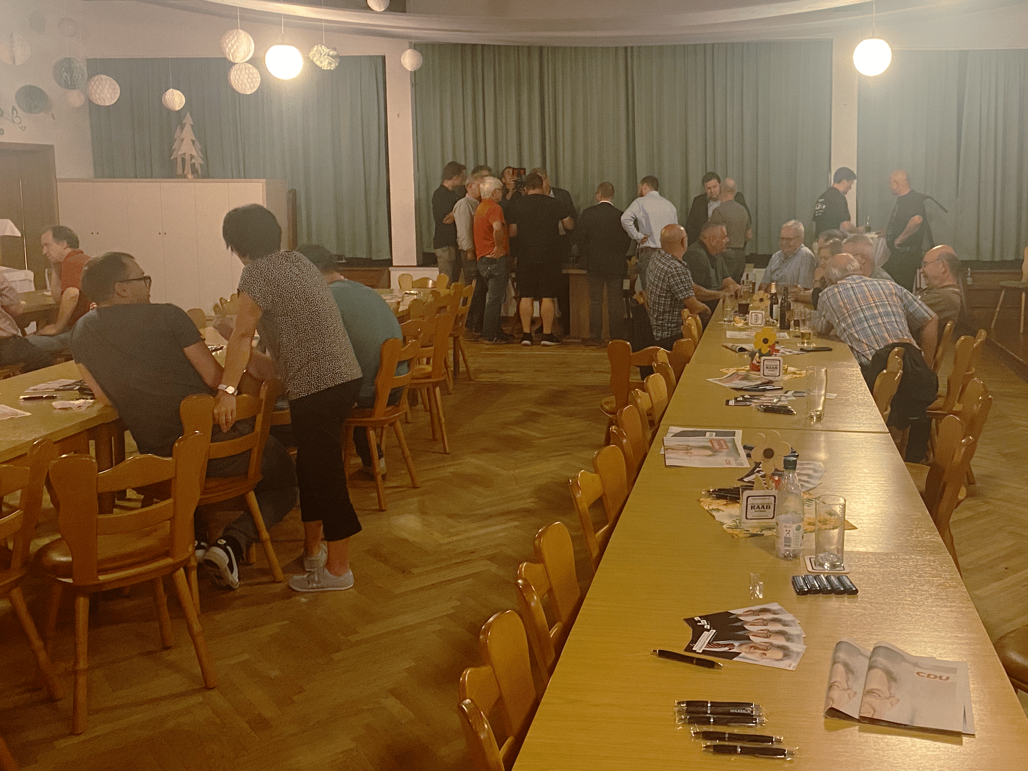 Ein Gastraum der sich langsam leer. Lange Tische, mit vereinzelt Gruppen, am anderen Ende des Raumes steht eine Gruppe Menschen.
