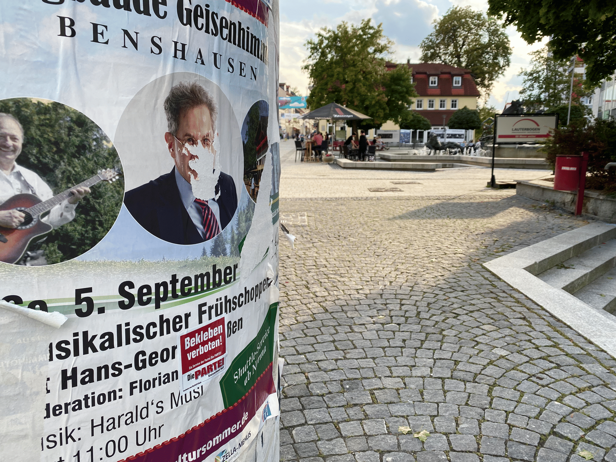 Links eine Litfaßsäule mit einem Plakat für ein Veranstaltung mit Maaßen, rechts der sonnendurhflutete Marktplatz.