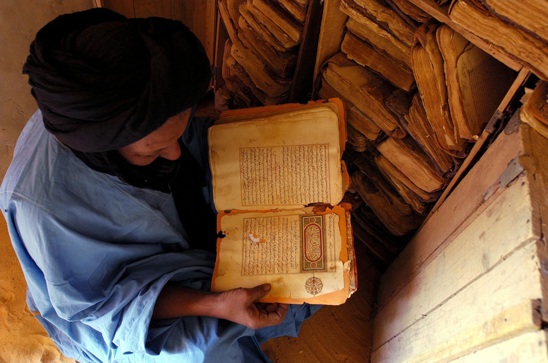 Ein bedounisch gekleidter Mann mit einem alten Buch in der Hand, von oben herab fotografiert, wodurch wir Kistenweise ähnliche Manuskripte um ihn sehen.