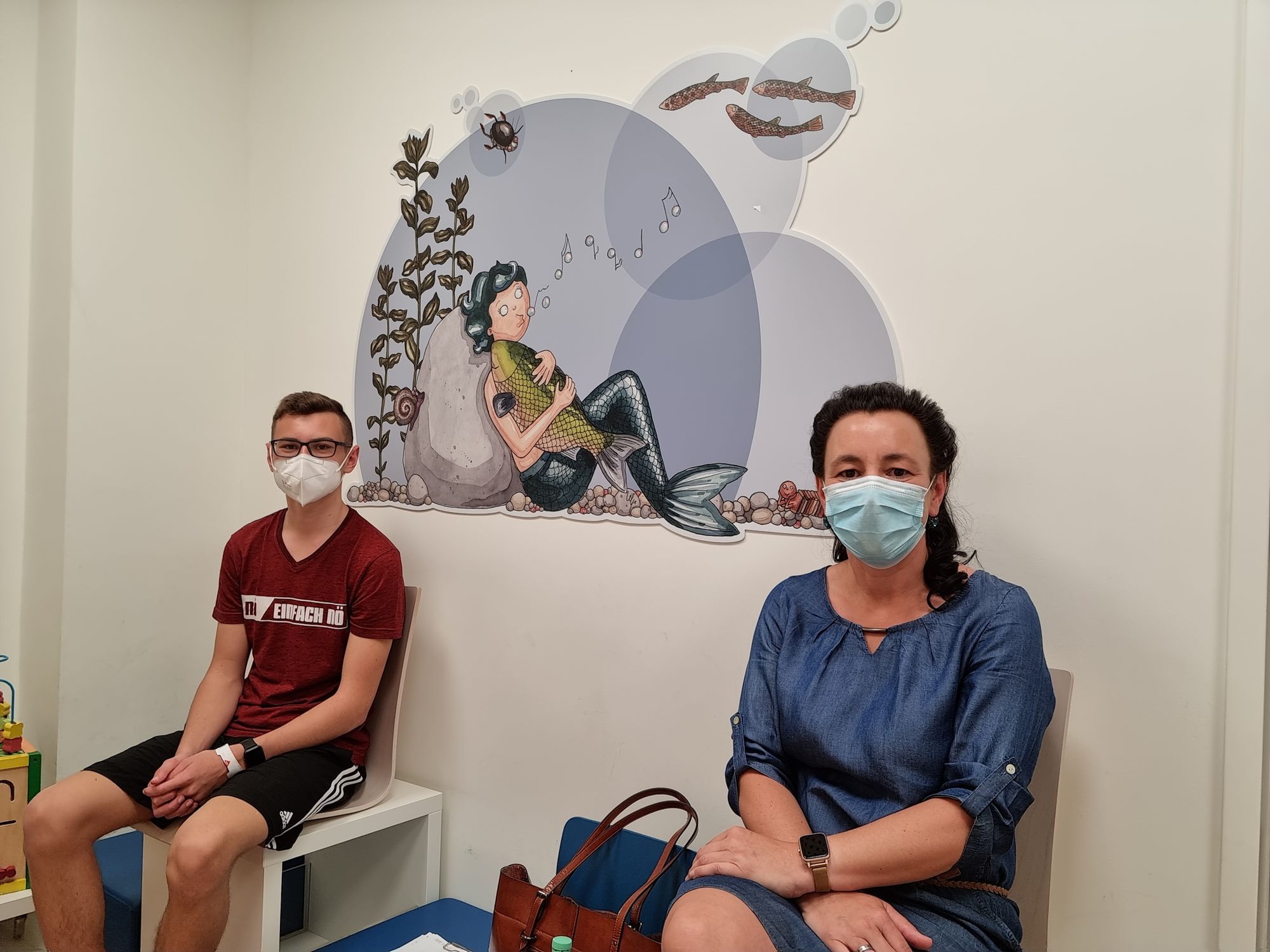 Links sitzt Felix, der Patient, auf einem weißen Hocker. Rechts sitzt seine Mutter. Sie tragen beide Masken. Im Hintergrund ist die Wand des Wartezimmers zu sehen, auf die eine Meerjungfrau aufgeklebt ist.