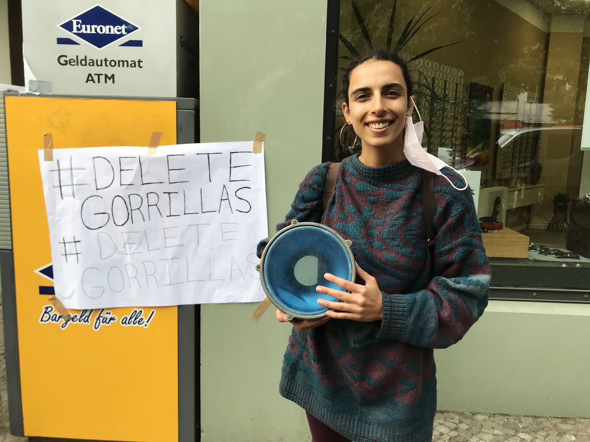 Eine Frau schaut und direkt an, ein Mundnasenschutz baumelt an ihrem linken Ohr. Sie hält eine Trommel. Hinter an einem Bankautomaten hängt ein Plakat mit der Aufschrift #deletegorillas