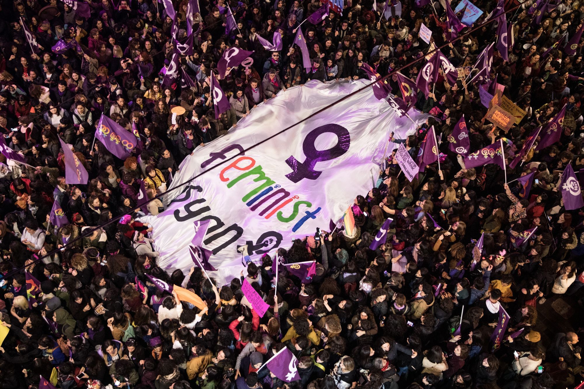 Eine Menschenmenge von oben herab, in ihrer ist ein großes weißes Banner mit der Aufschrift "Feminist" und dem Symbol für Weiblich in Violett.