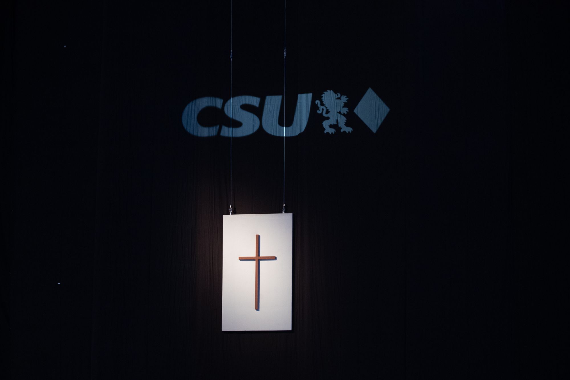 Ein Holzkreuz auf weißem Hintergrund ist mit Seilen von oben abgespannt und angestrahlt. Im hintergrund ist ein schwarzer Vorhang mit dem CSU Parteilogo.