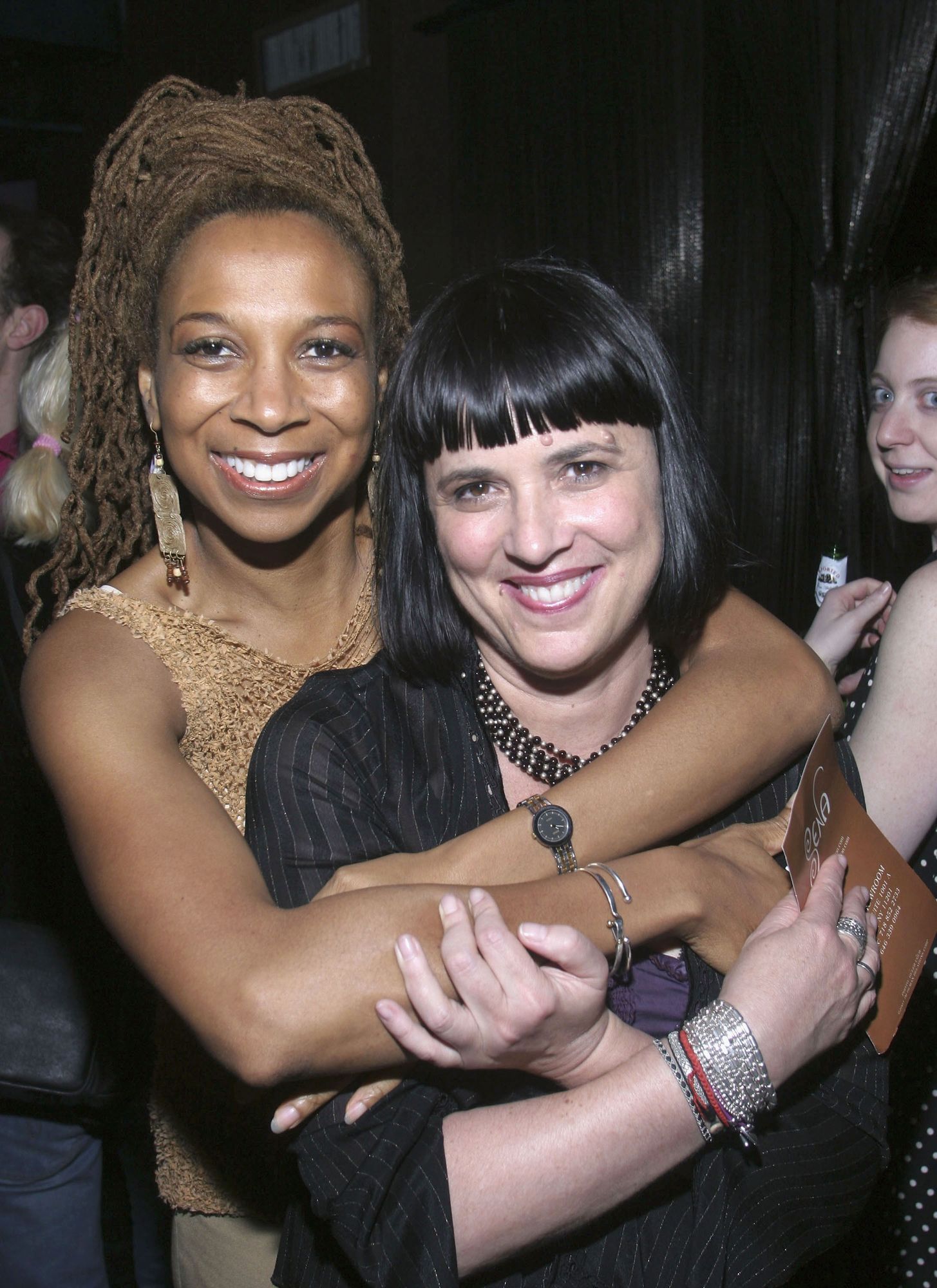 Crenshaw, eine schwarze Frau mit hochgesteckten Haaren mit Ohrenringen und Kleid, umarmt eine andere Frau, Eve Ensler, die weiß mit schwarzen Haaren ist. Beide Lächeln in die Kamera.