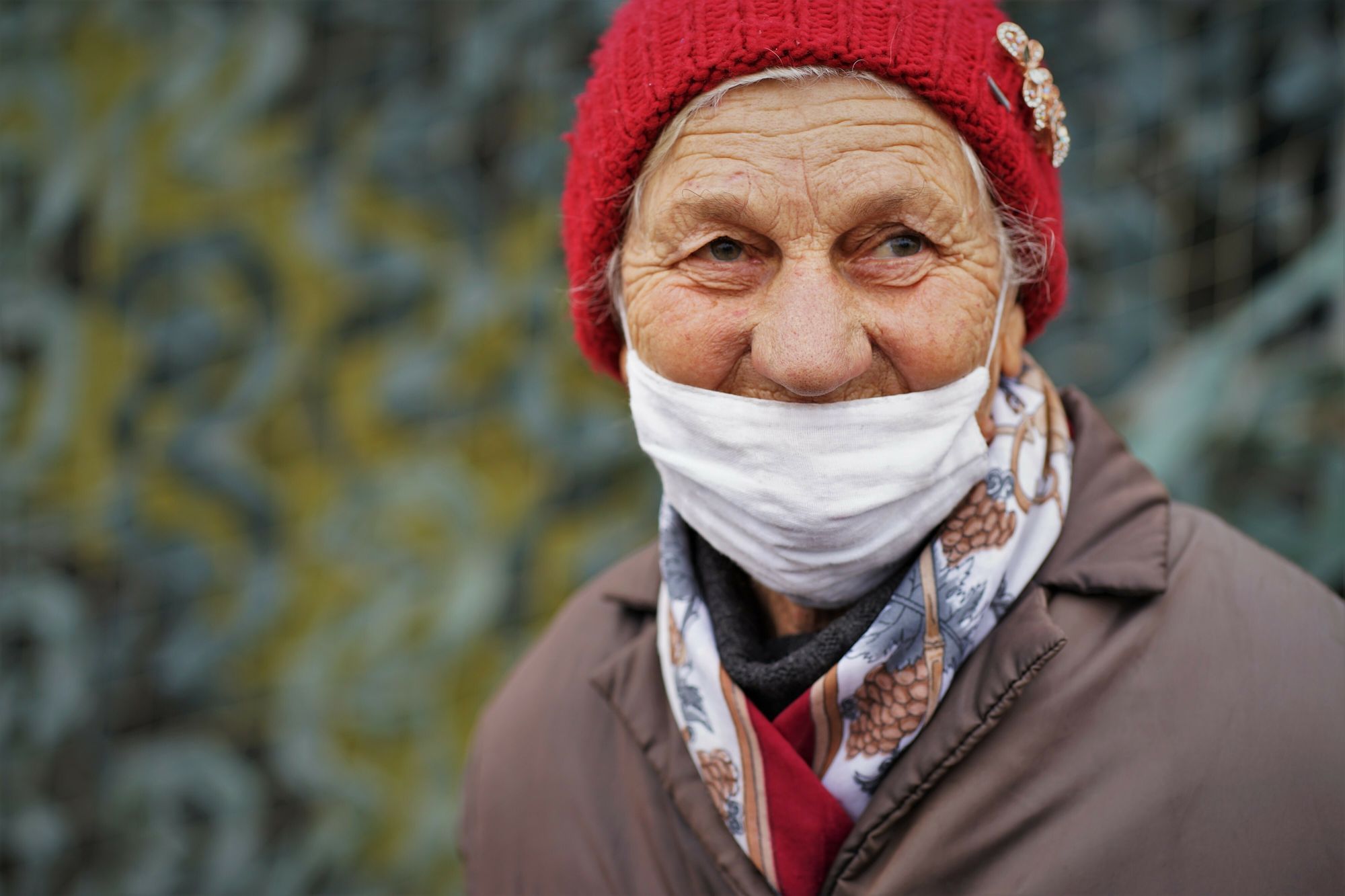 Eine alte Frau mit roter Strickmütze und dem Mundnasenschutz knapp under der Nase.