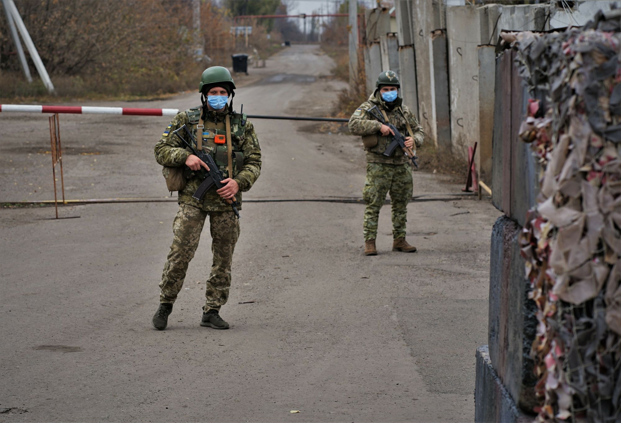 Eine Straße in schlechtem Zustand, mit einem Schlagbaum und zwei Soldaten davor. Sie tragen Helm, Mundnasenschutz und Kalaschnikow.