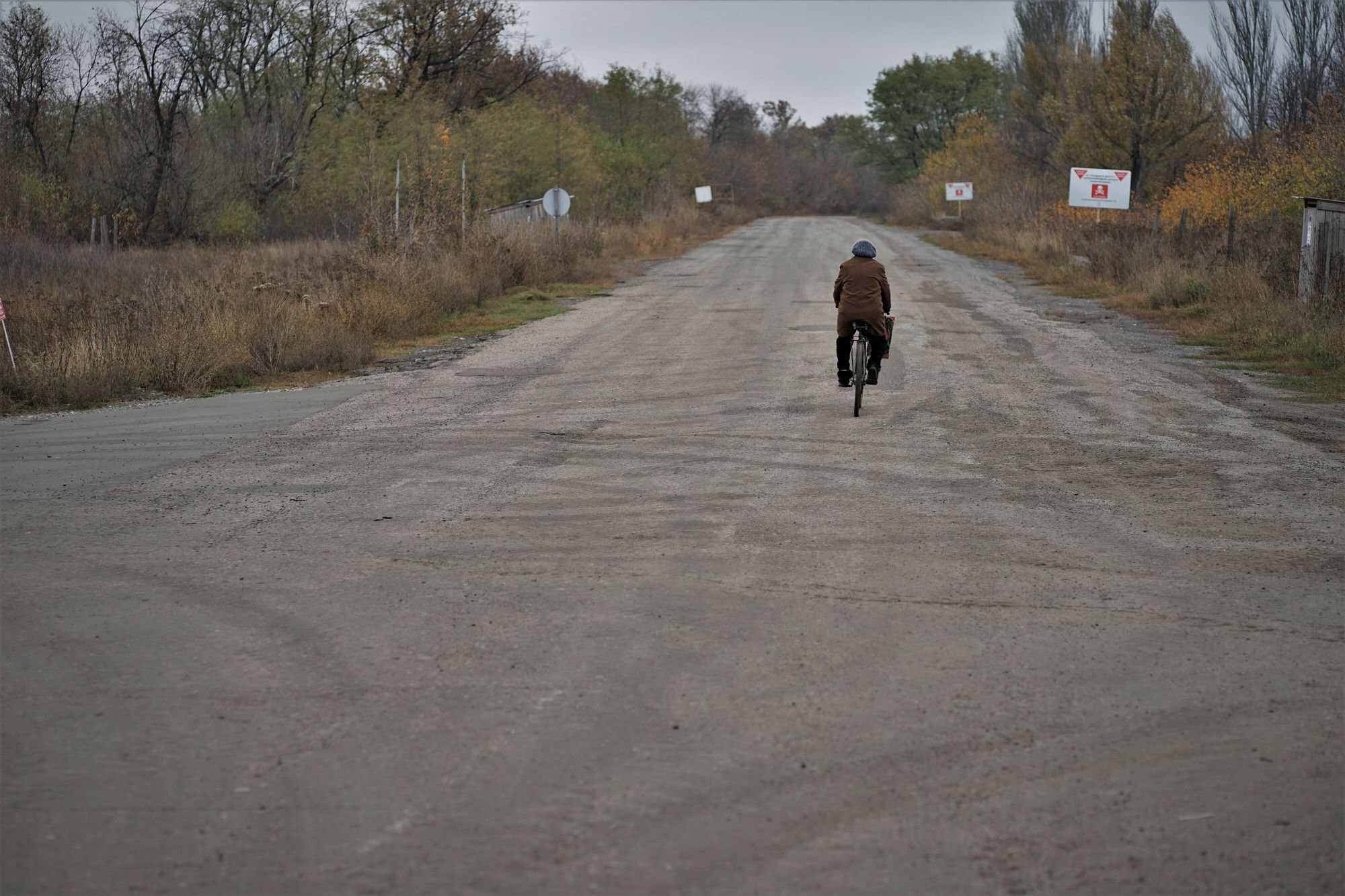 Eine Straße in desolatem Zustand auf der eine weibliche gelesene Person mit dem Fahrrad fährt. Die Umgebung macht einen gedeckten, drückenden Eindruck. Links und rechts der Straße sind Schilder mit Landminenwarnungen.