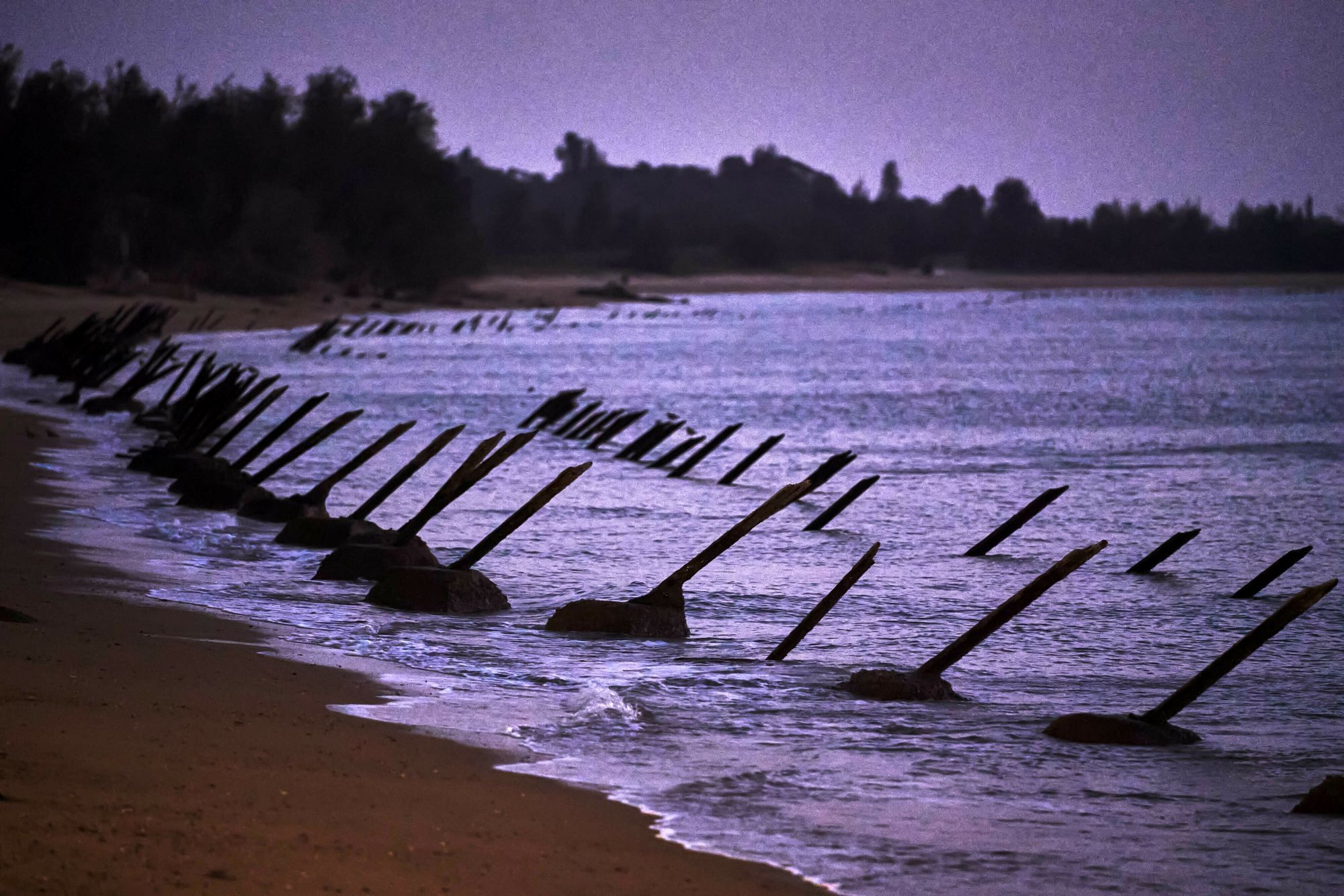 Abendstimmung, ein Sandstrand. Aus der Brandung stechen schwarze Balken hervor.