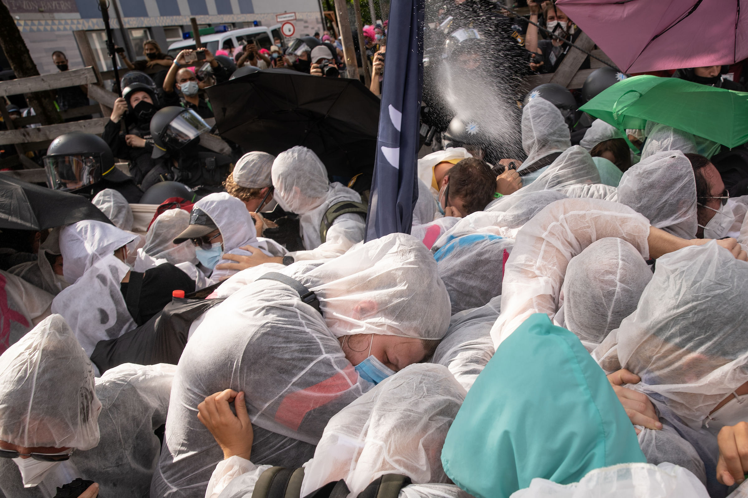 Eine Gruppe von Klimaaktivistis in weißen Malerschutzanzügen presst sich aneinander während aus einer schwarzen Masse von Polizist:innen herraus Pfefferspray eingesetzt wird.