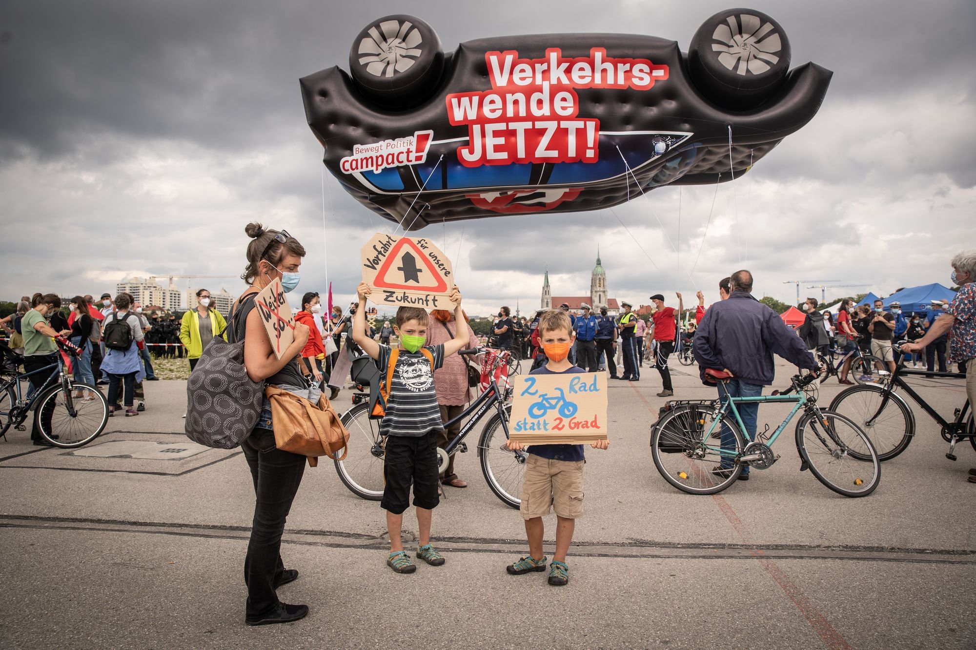 Eine weibliche gelesene Person, vermutlich ein Elternteil, steht mit zwei zwei männlich gelesenen Kindern und Schildern auf einer Demonstration für die Verkehrswende.