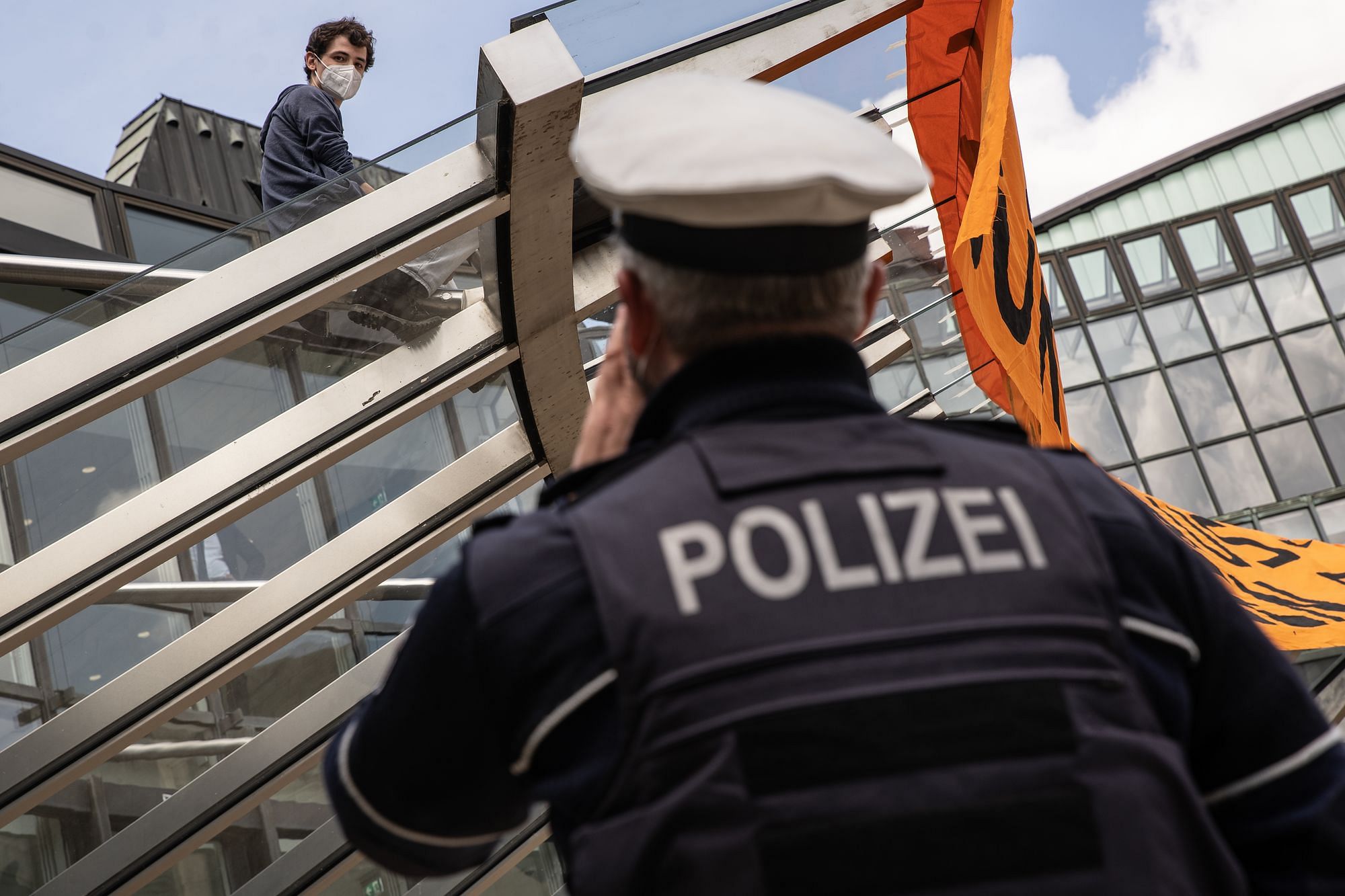 Ein Klimaktivisti mit Banner sitzt auf dem gläsernen Vordach des Landtags und schaut auf eine:n Polizist:in herab die mit Rücken unscharf im Vordergrund steht.