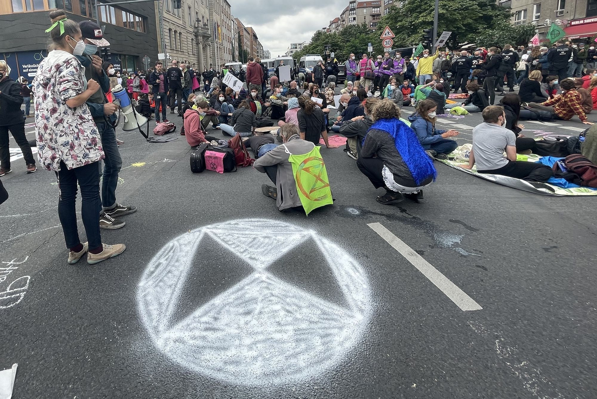 Klimaaktivisti sitzen auf der Straße, friedlich zusammen um diese zu blockieren. Im Vordergrund ist das Logo von Extinction Rebellion aufgesprüht.
