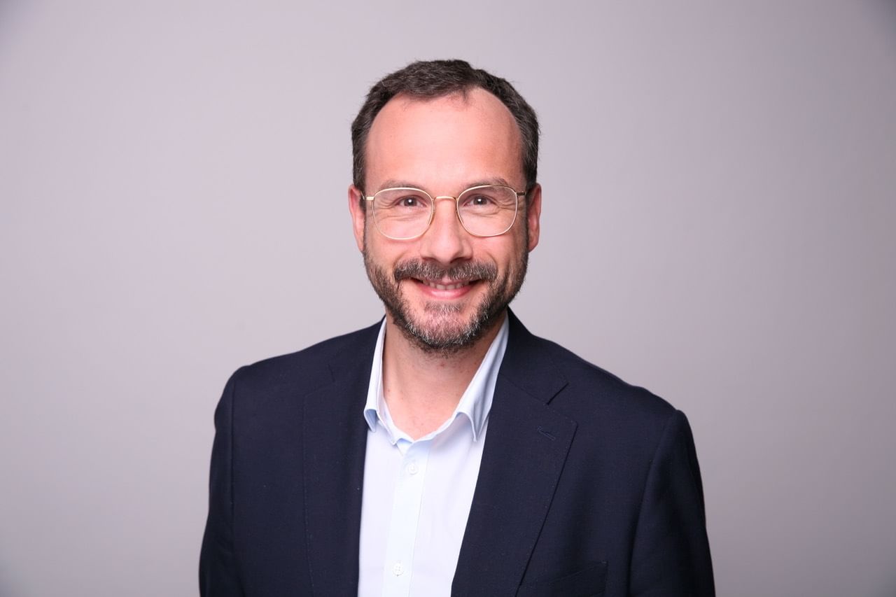 Ein Mann mittleren Alters im Sacko, mit Brille und fliehender Stirn.