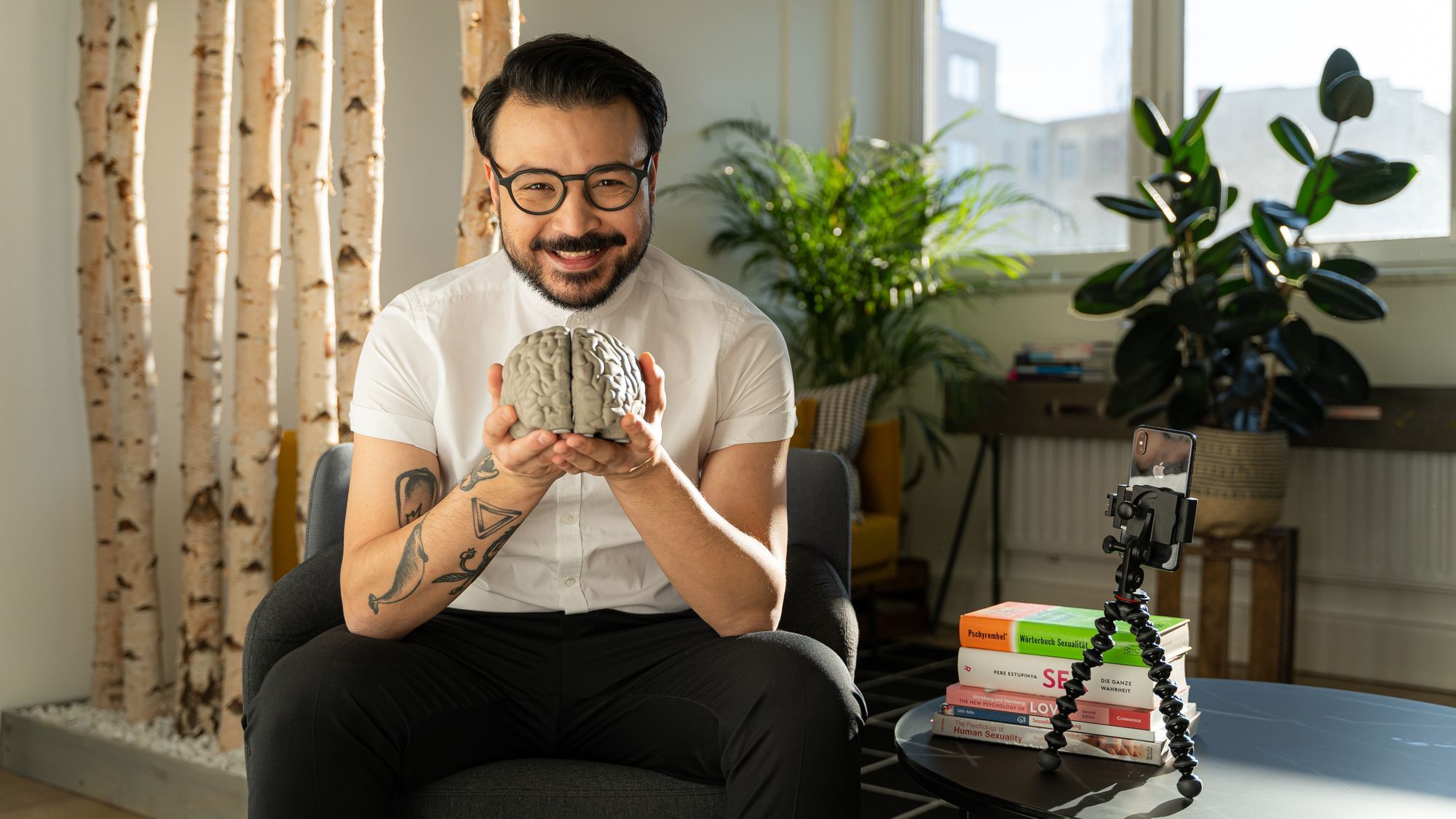 Ein Mann in seien frühen 30ern sitzt vor einer moderen Wohnlandschaft. Er hält ein Gehirn in der Hand und lächelt in die Kamera. Neben ihm liegen sind Bücher über Sexualitäten.