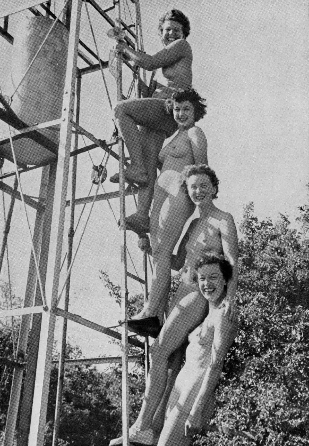 Vier nackte Frauen schauen auf uns herab von der Leiter einer Art Metall- Wasserturm.