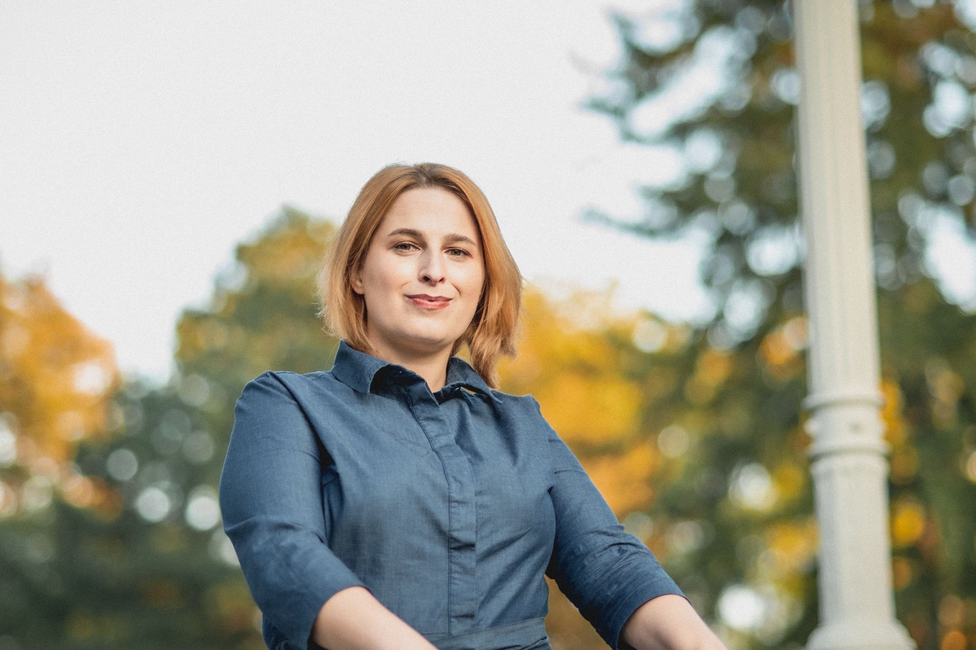 Eine weiblich gelesene Person mit Blond-Rotem Haar, in einem blauen Oberteil. Sie blickt herrausfordernd in die Kamera.