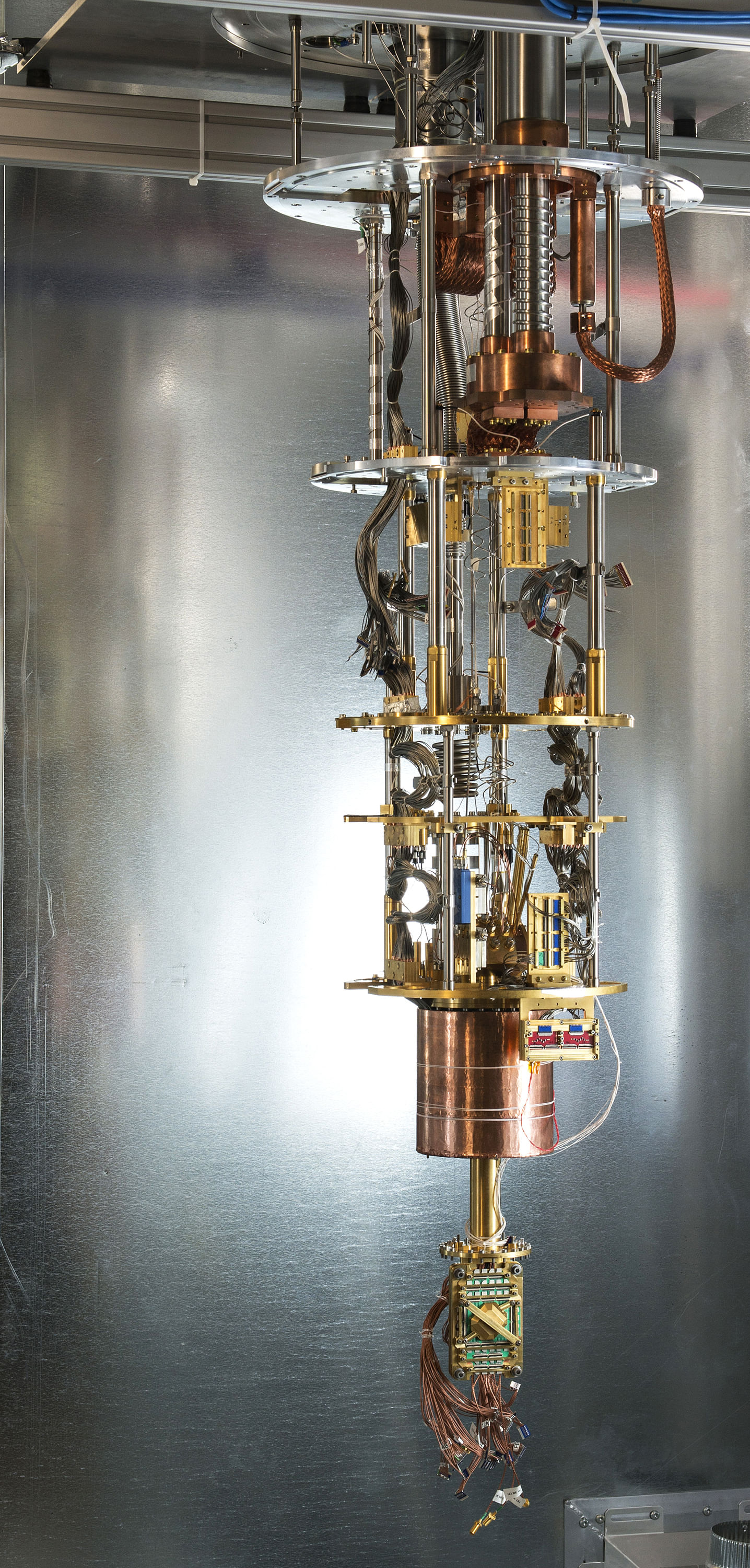 Wie ein länglicher Kronleuchter sieht das Innenleben des Quantencomputer aus, aufgehängt an der Decke. Fünf Scheiben aus goldenem und silbernem Metall bilden die Etagen des Kronleuchters, dicke Stränge von Kupferdrähten winden sich von oben herab bis unten zur Spitze.