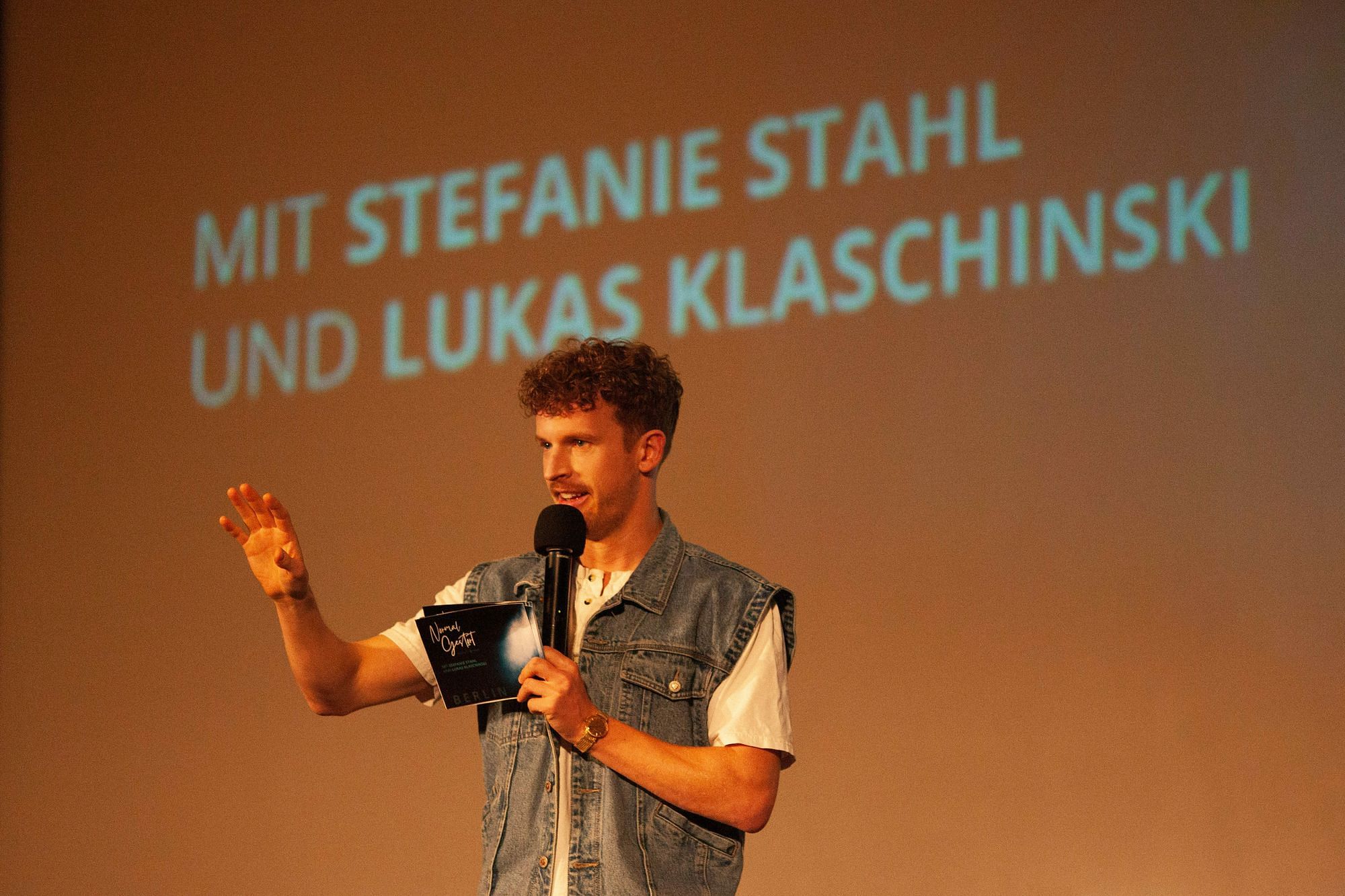 Wir sehen den Moderator Lukas Klaschinski, der ins Mikro spricht und mit einer Handbewegung etwas andeutet