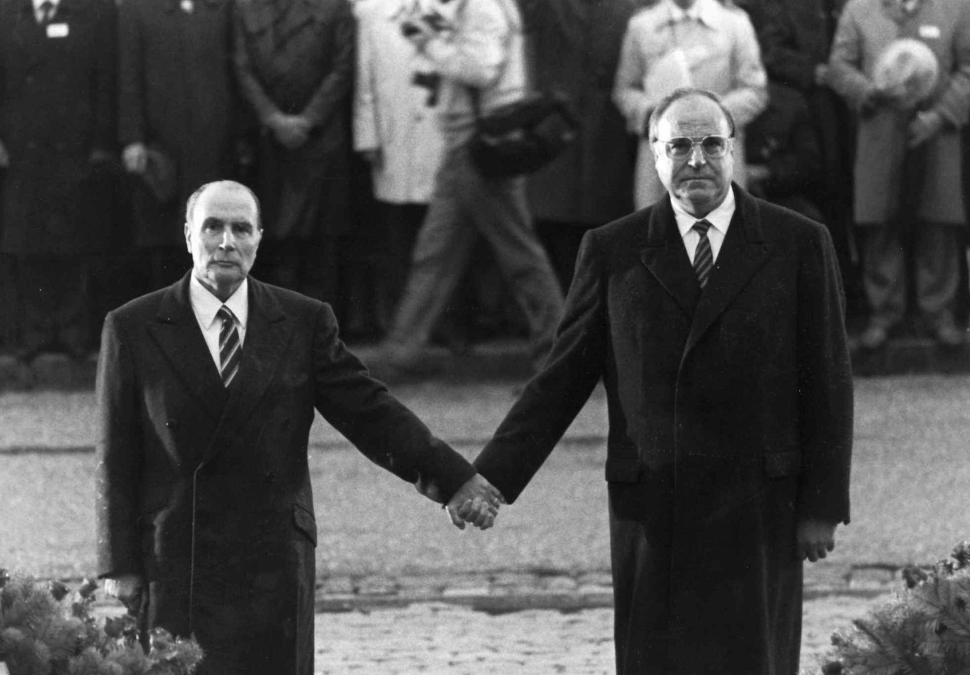 Helmut Kohl und Francois Mitterand halten sich in Verdun an den Händen. Im Hintergrund sieht man mehrere Menschen, die in etwas Abstand zu den beiden stehen. Vor den beiden befinden sich Blumenkränze.