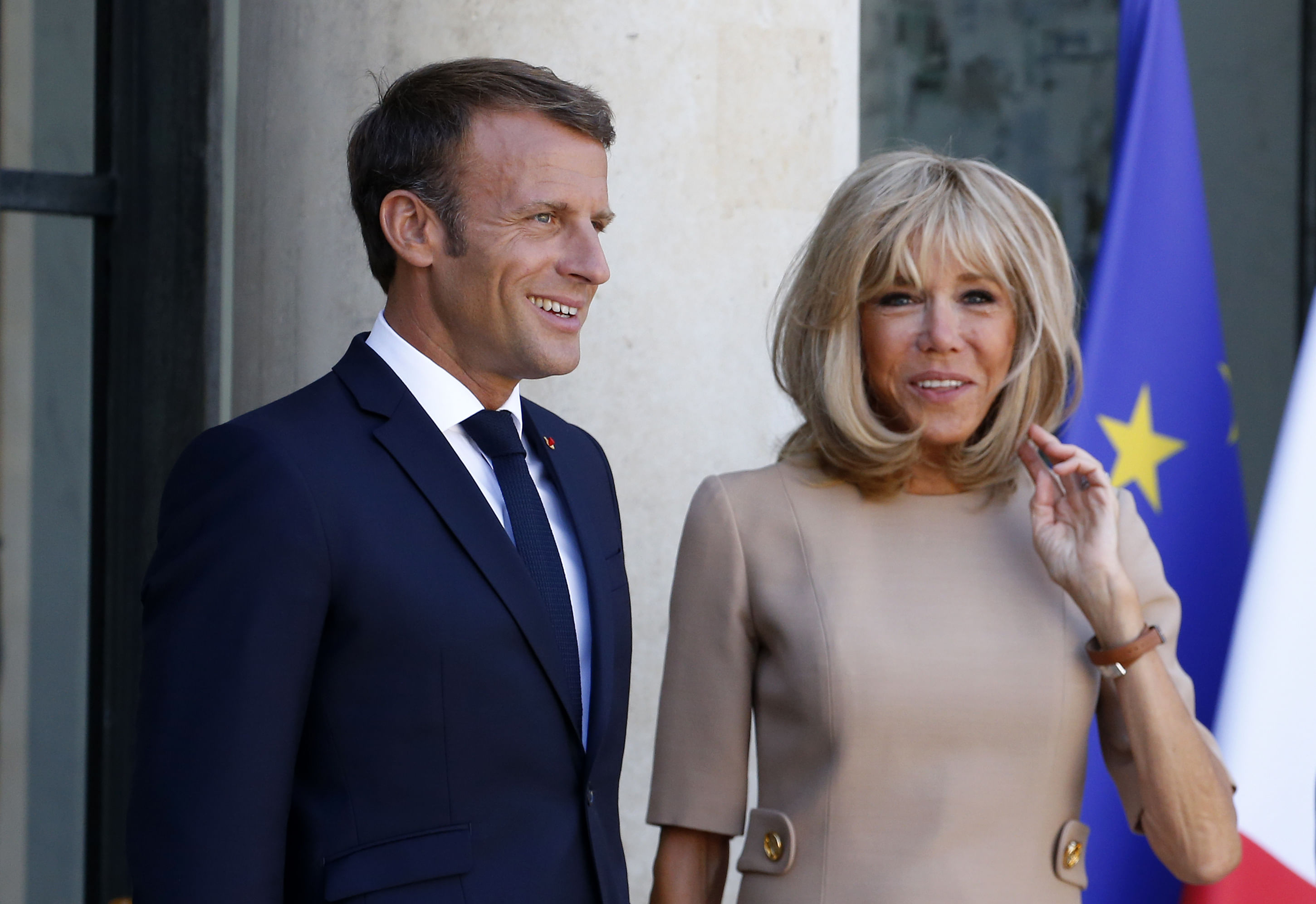Emmanuel Macron und seine Frau Brigitte. Er trägt einen Anzug und sie ein beigefarbenes Kleid. Im Hintergrund sieht man die europäische und die französische Flagge.