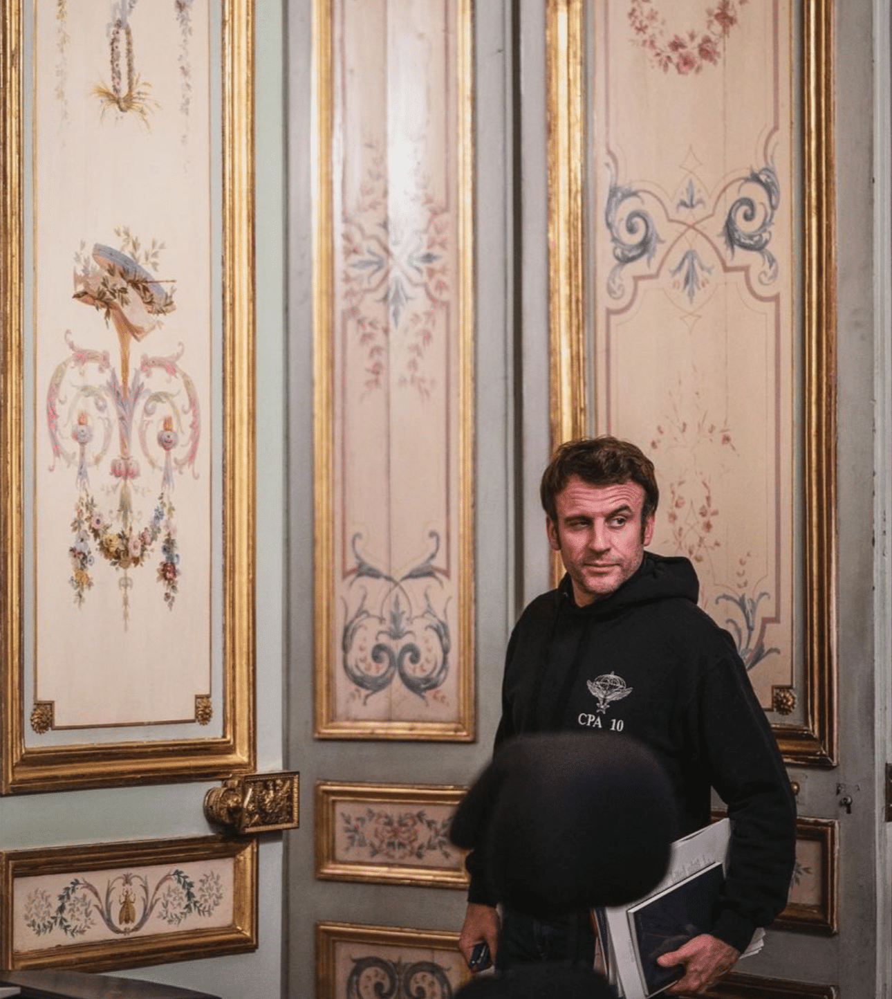 Macron kneift ein Auge zu und blickt an der Kamera vorbei, unter dem Arm trägt er einen Stapel Akten. Er trägt einen Kapuzenpullover.