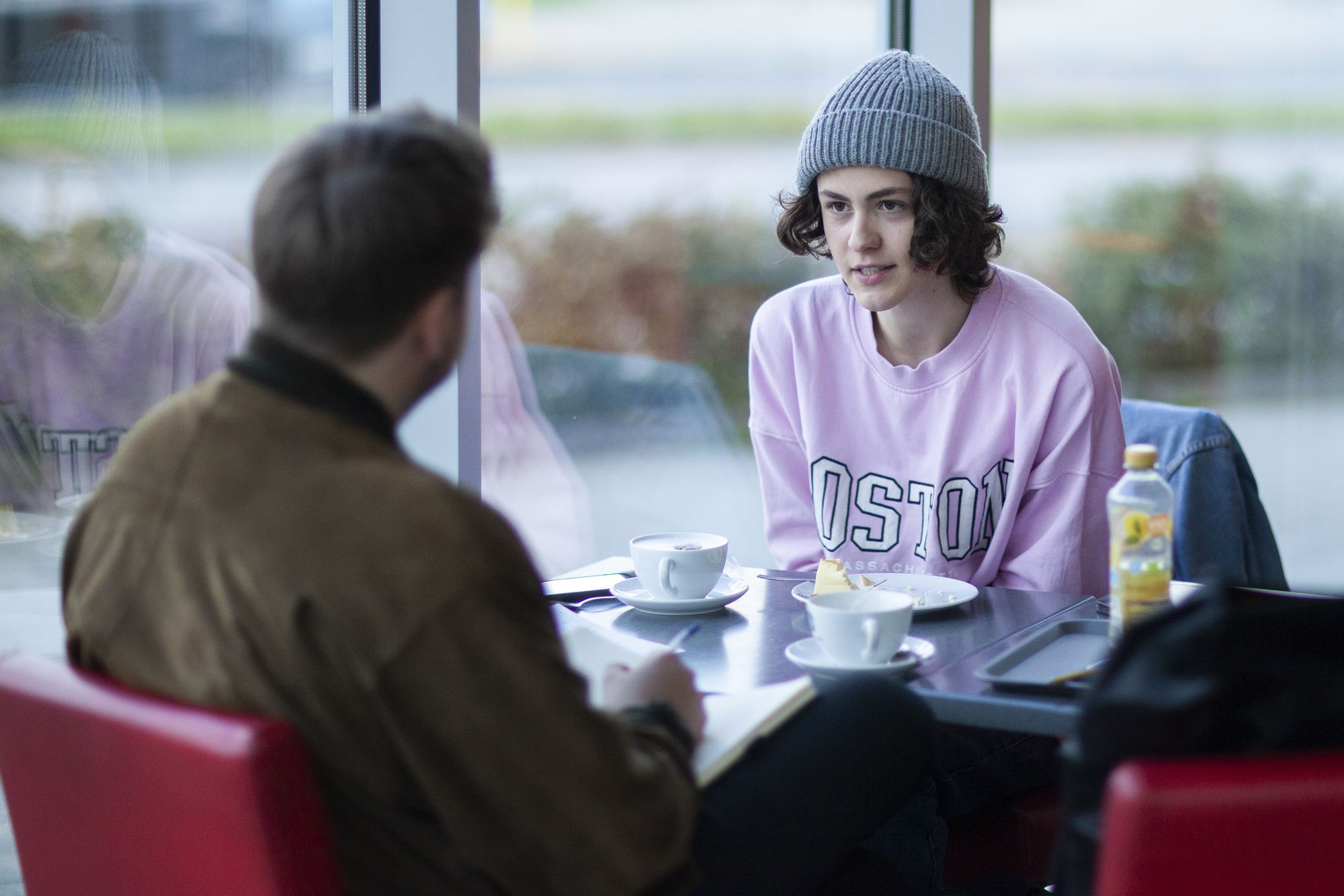 Gianni und Bent sitzen sich einem Café gegenüber und unterhalten sich. Bent trägt eine braune Lederjacke.