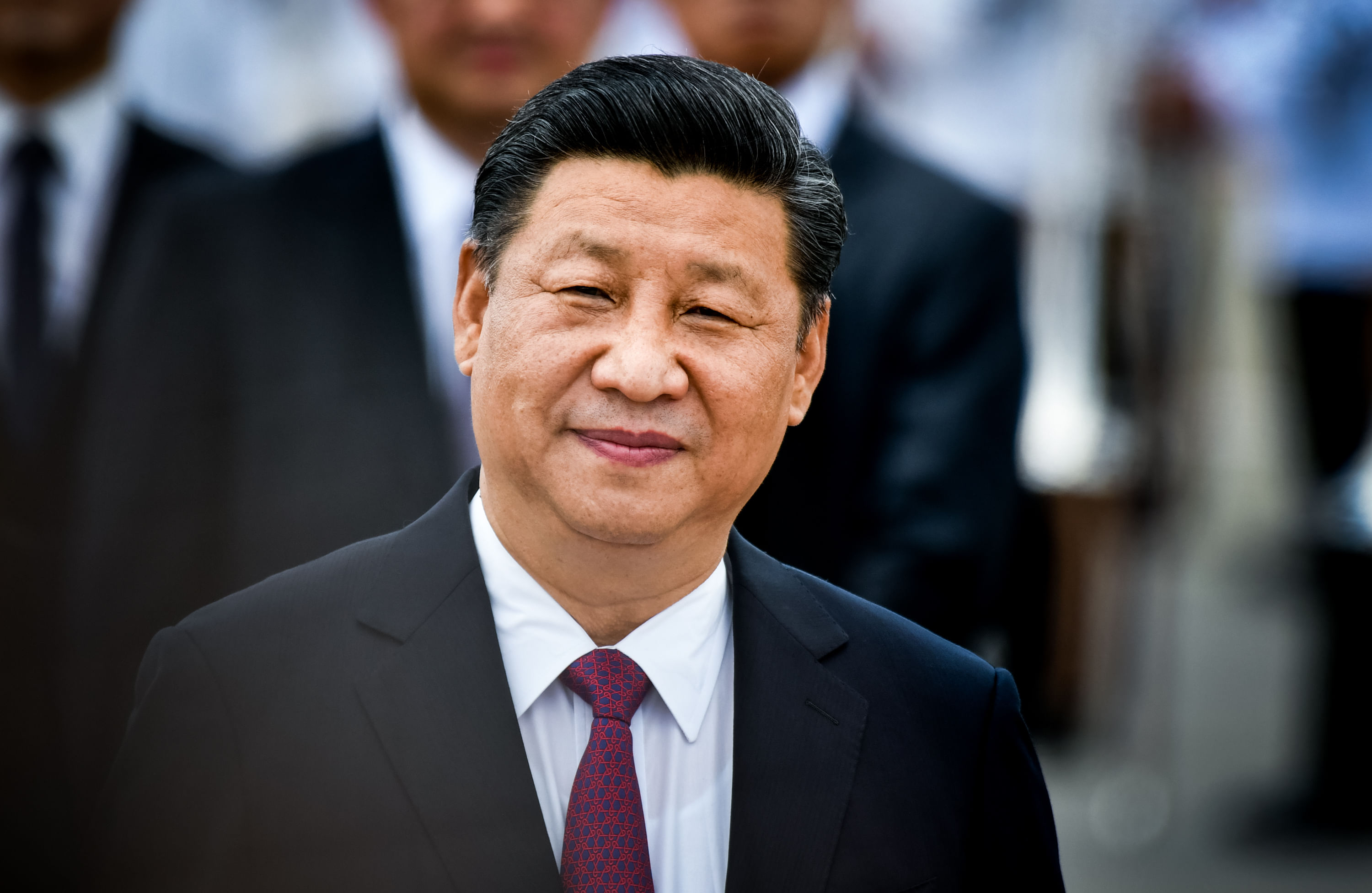 Xi Jinping trägt einen Anzug und lächelt in die Kamera. Im Hintergrund sind mehrere Personen im Anzug zu erkennen.