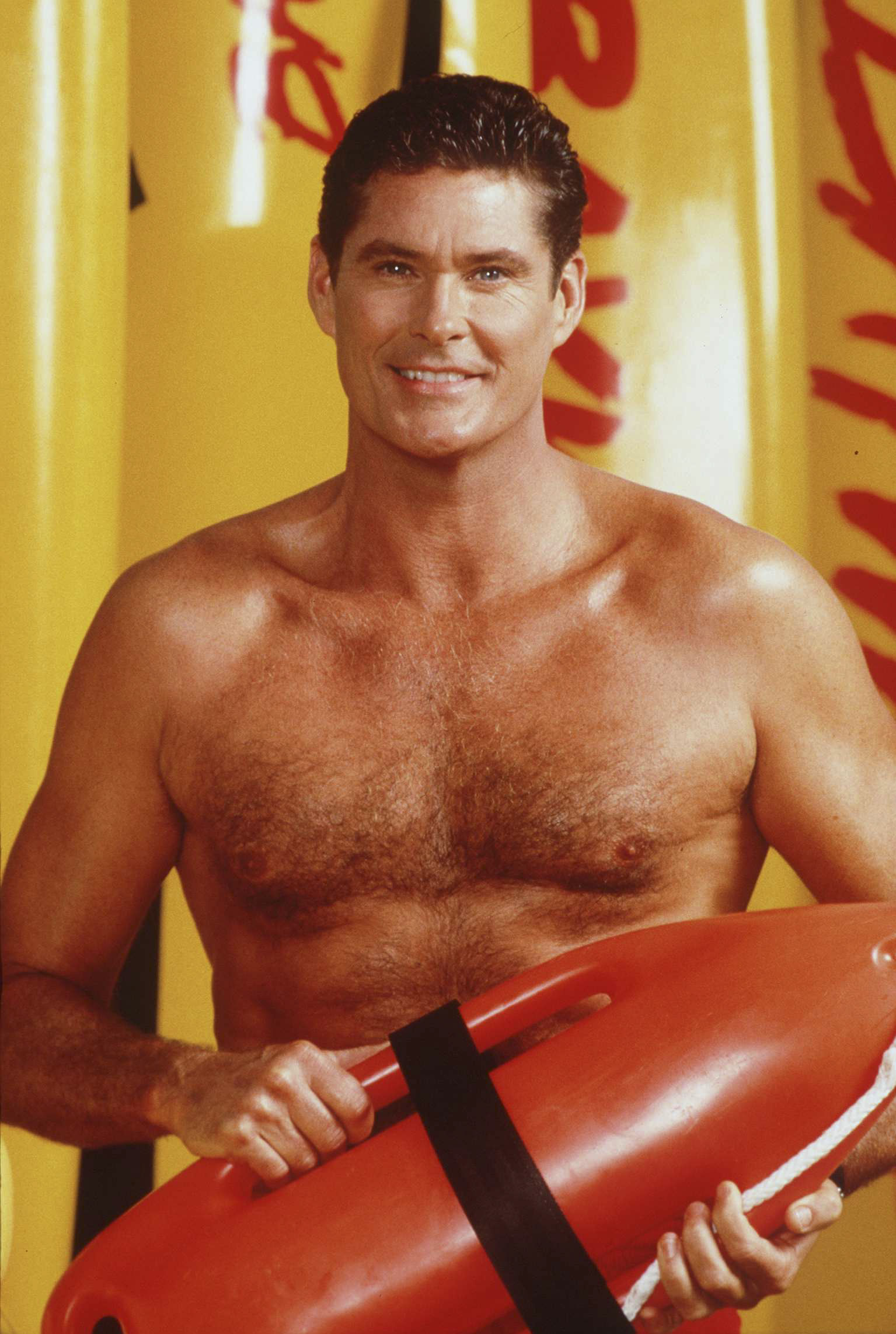 David Hasselhoff lächelt oberkörperfrei in die Kamera. Er trägt die bekannte orangefarbene Schwimmboje aus Baywatch.