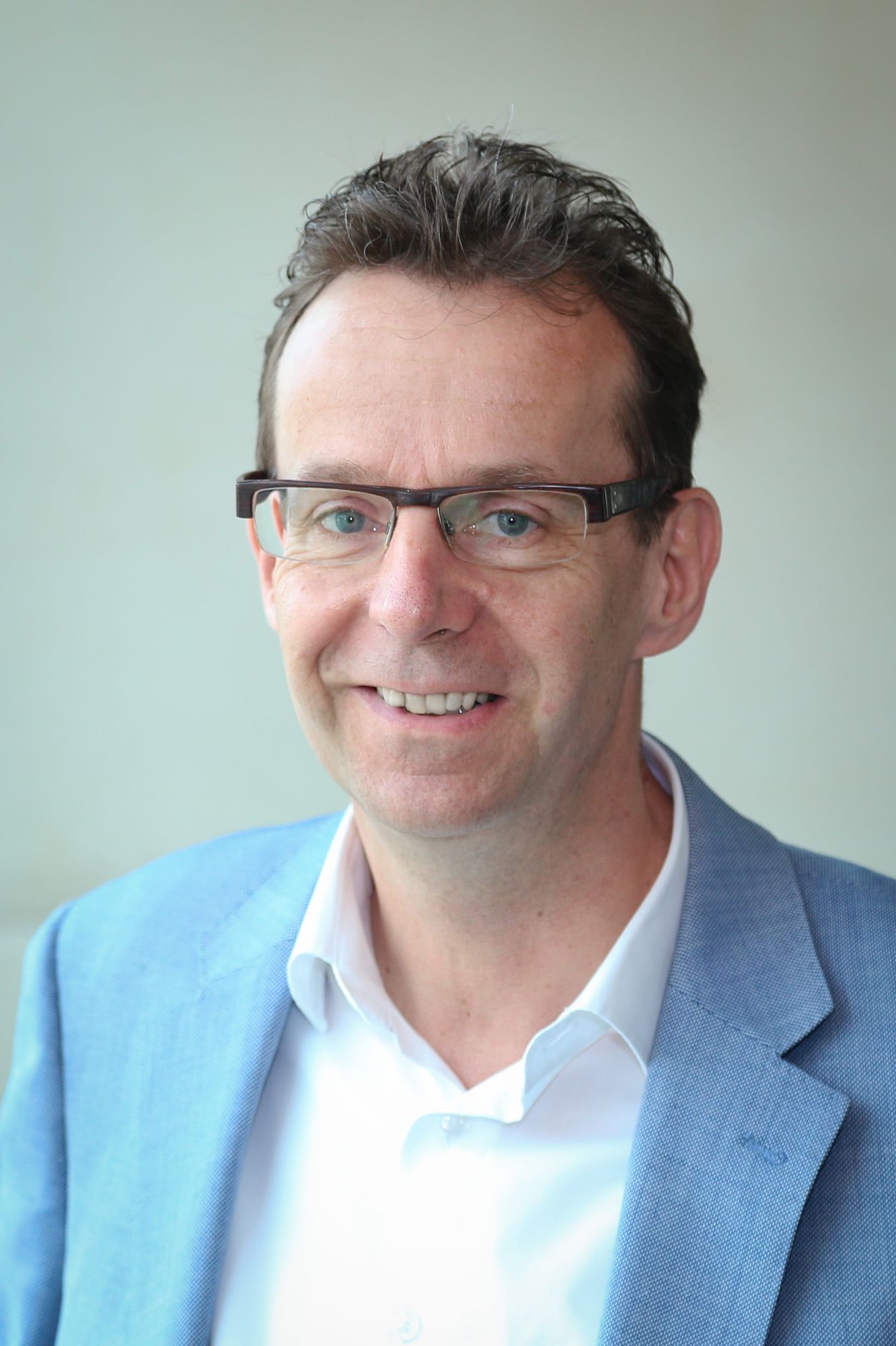 Man sieht einen Mann in einer Pirtraiaufnahme, also das Gesicht, den Hals, die Brust. Der Mann trägt ein weißes Hemd, ein hellblaues Jacket und eine Brille, er lächelt.