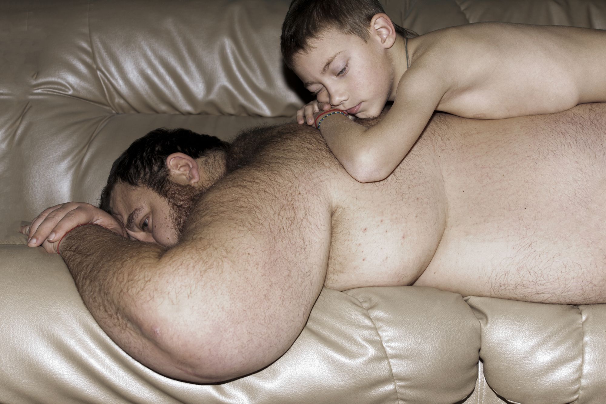 Wir sehen ein Kind, das sich auf den Rücken seines Vaters legt, der auf der Couch liegt. Beide sind nackt.