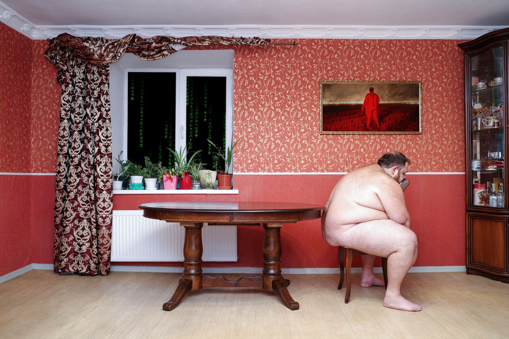 Ein Mann sitzt in einem roten Zimmer nackt auf einem Hocker.